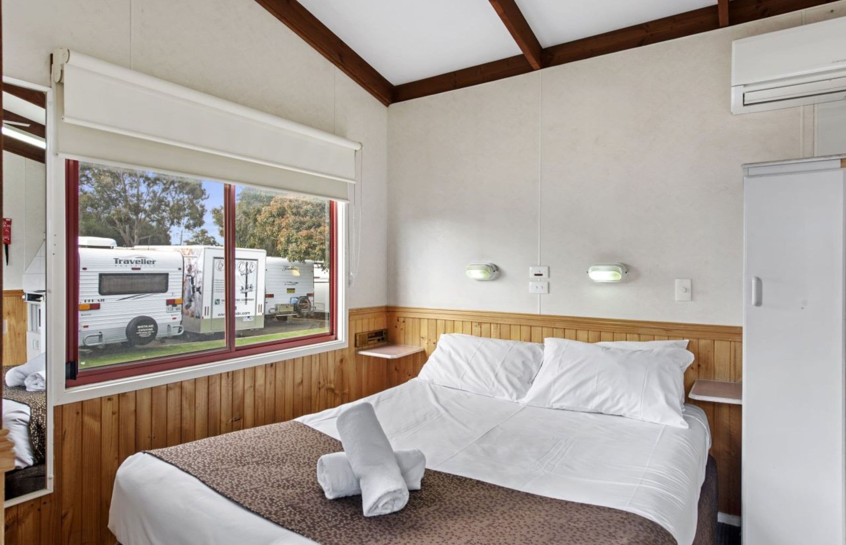 Un dormitorio con una cama grande, una ventana que da a vehículos recreativos, paredes de madera y un armario blanco.