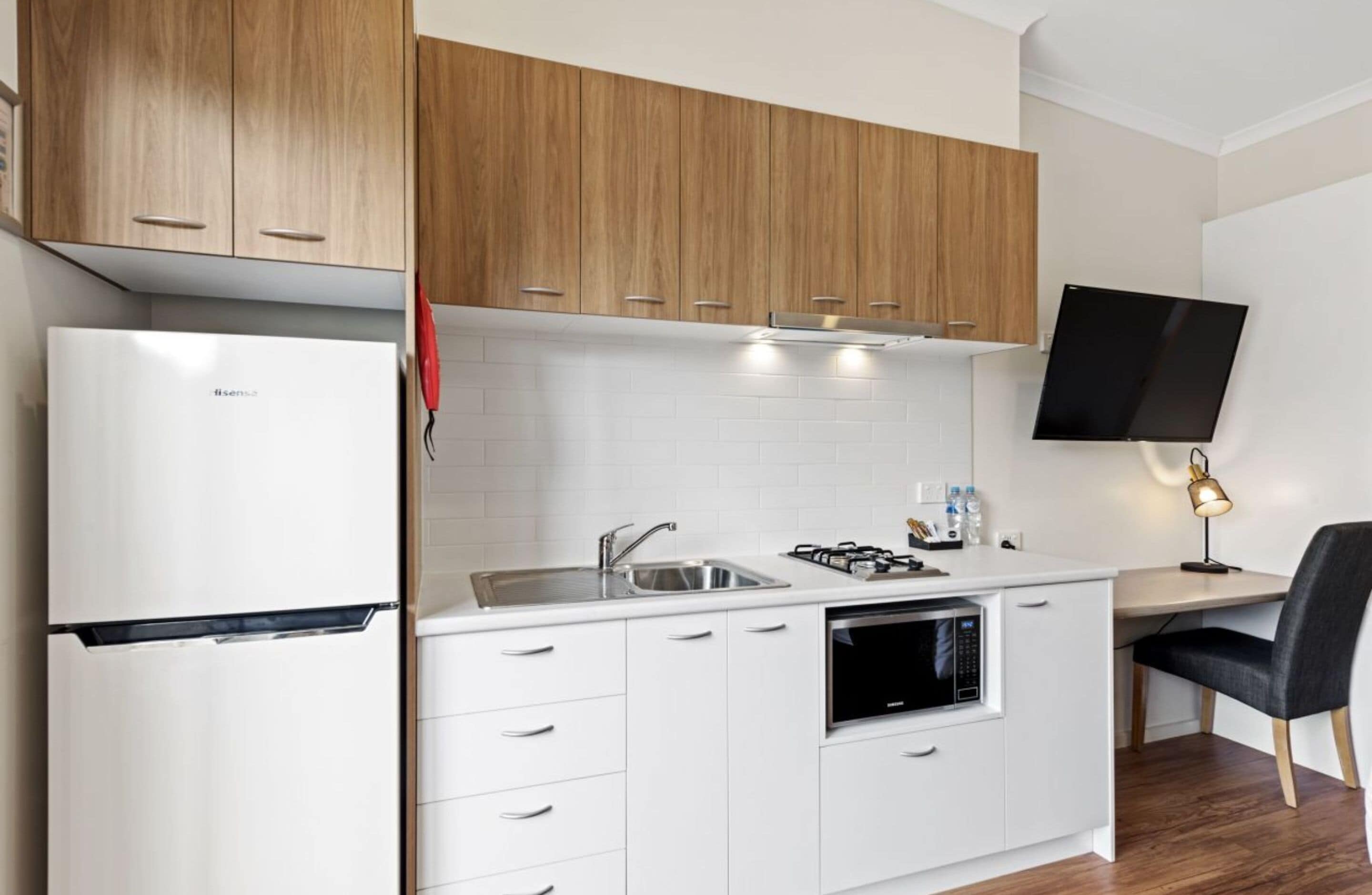 Cocina compacta con armarios blancos, microondas integrado, lavamanos y televisor de pantalla plana montado en la pared.