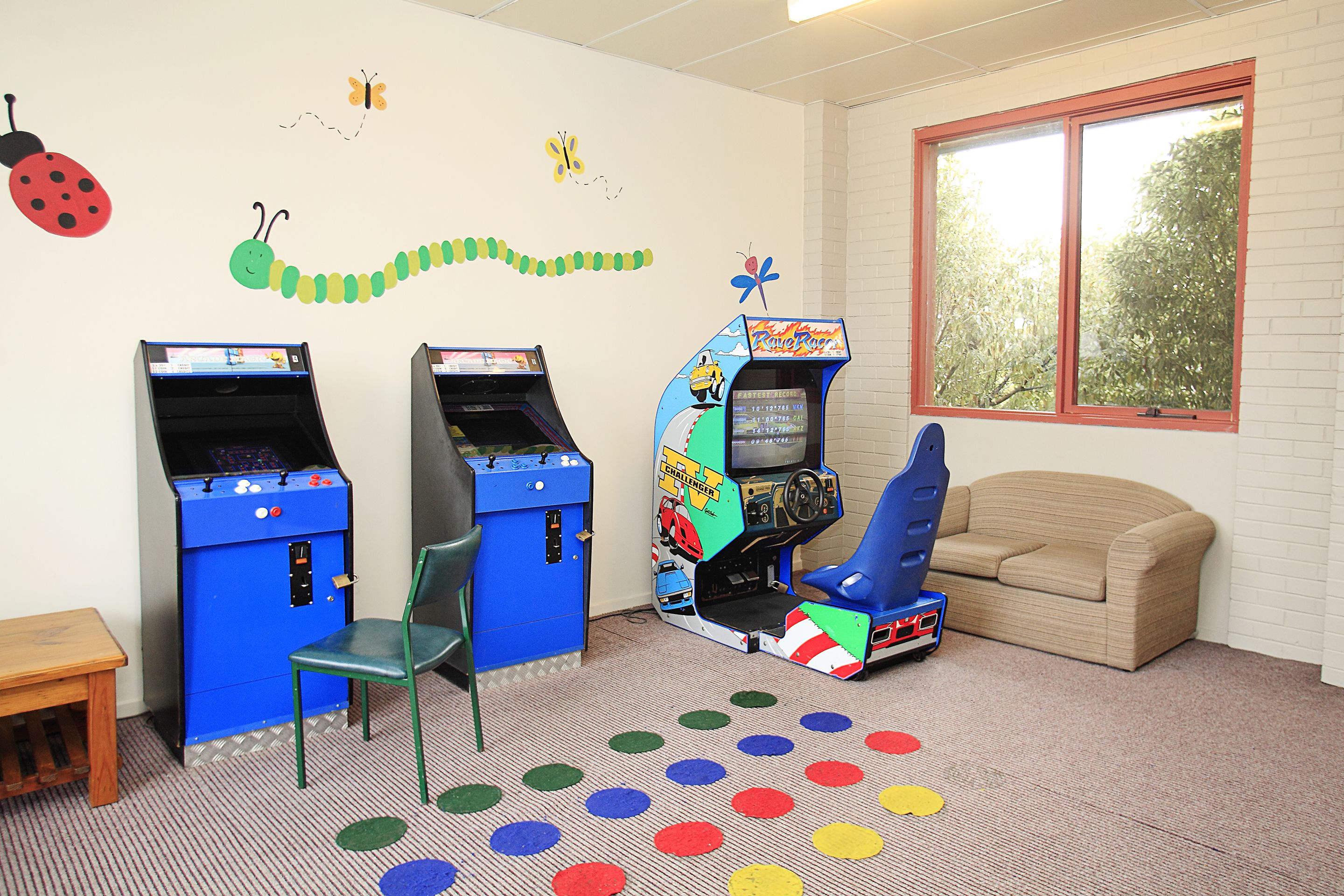 Una habitación con dos máquinas de videojuegos, un sillón azul, una mesita y un ventanal con vistas a árboles.