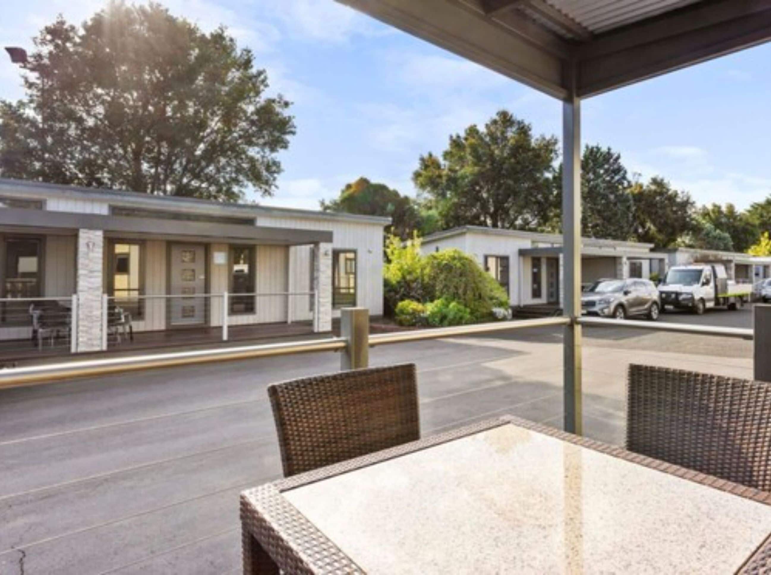 A covered outdoor seating area with wicker furniture, a building with a flat roof, and parked cars in the background.