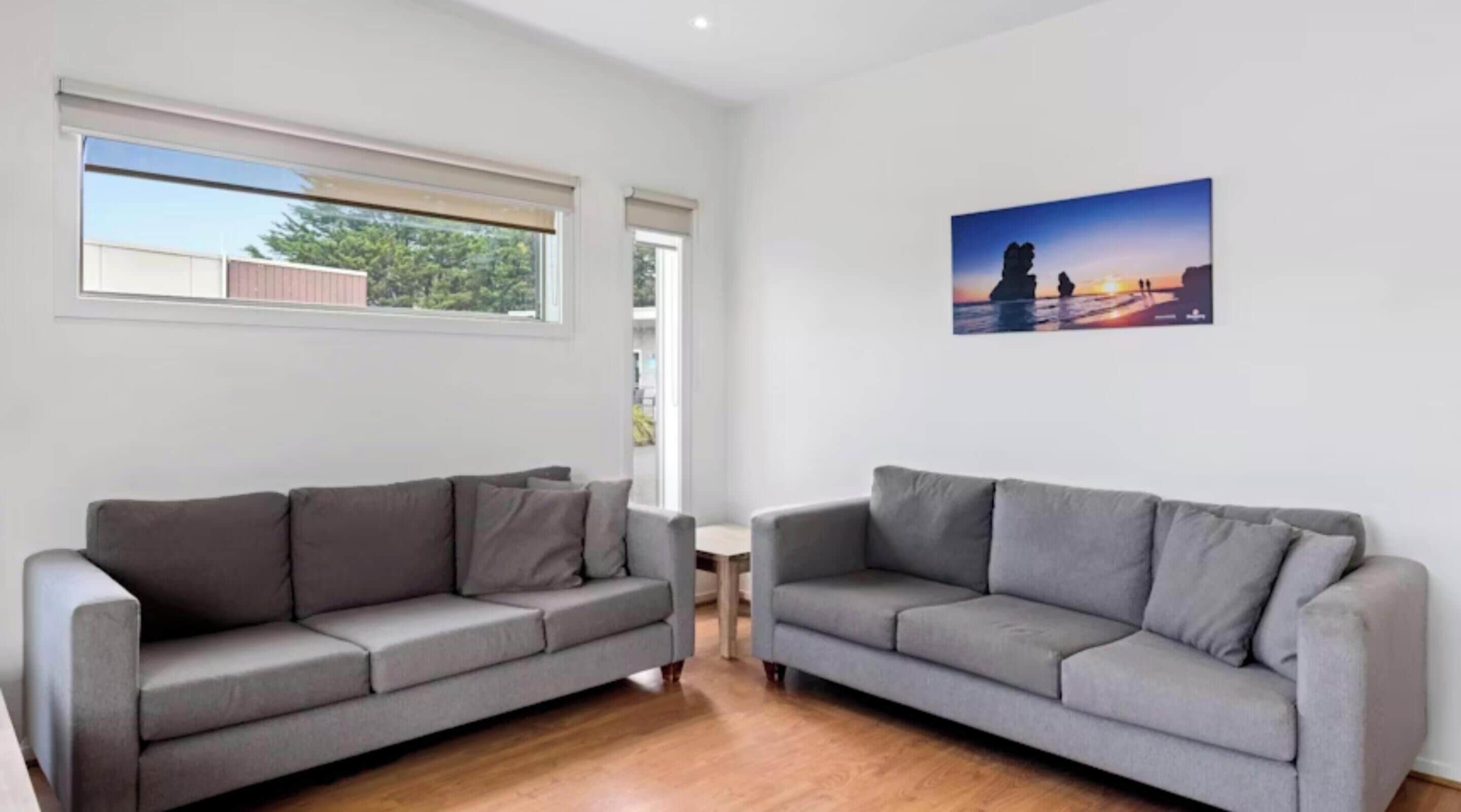 A modern living room with two grey sofas, a small round table, and a large window with a view of greenery.