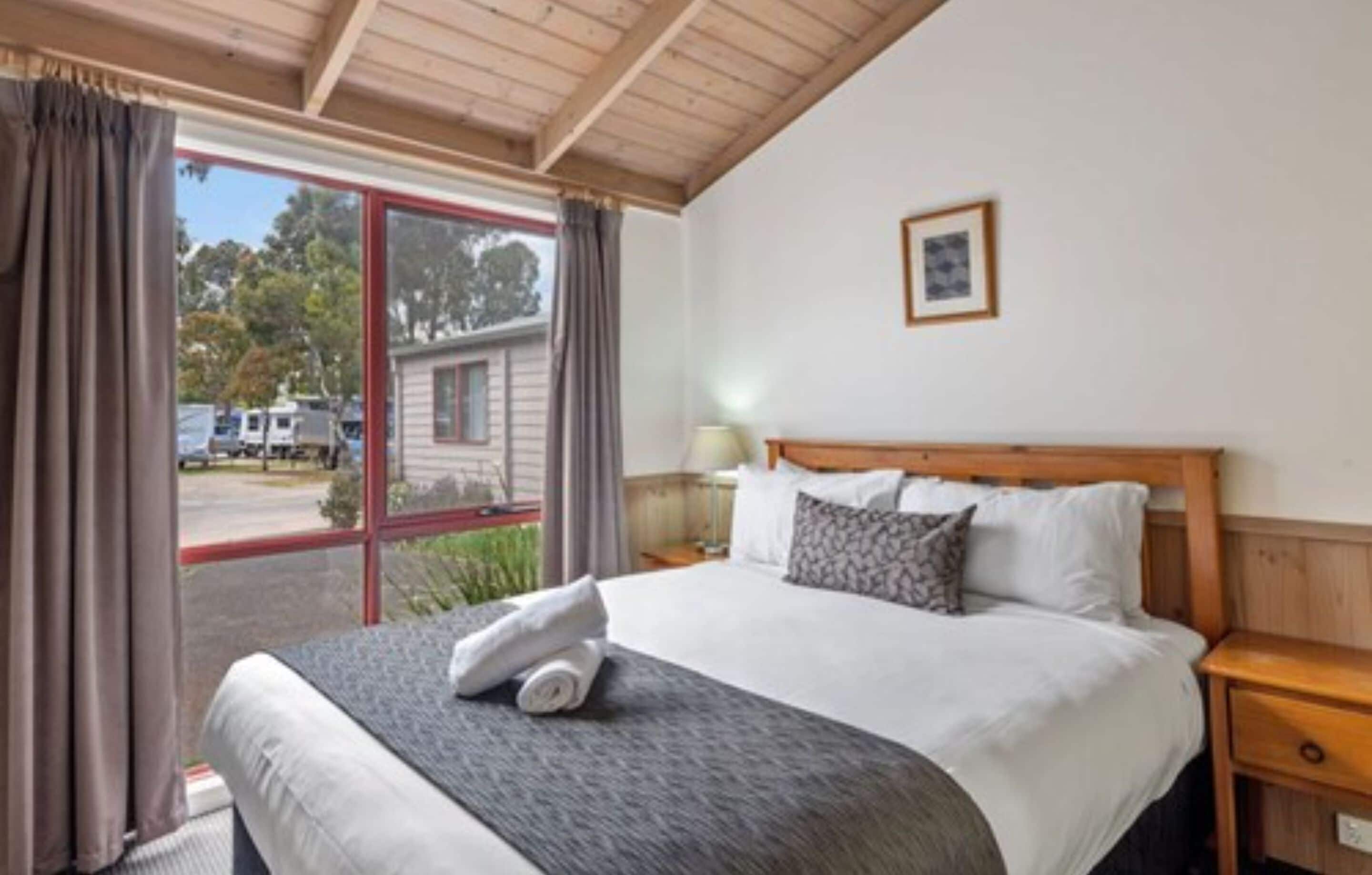 Una habitación de hotel con una cama grande, cabecera de madera, una mesita de noche, una lámpara y una ventana con cortinas.