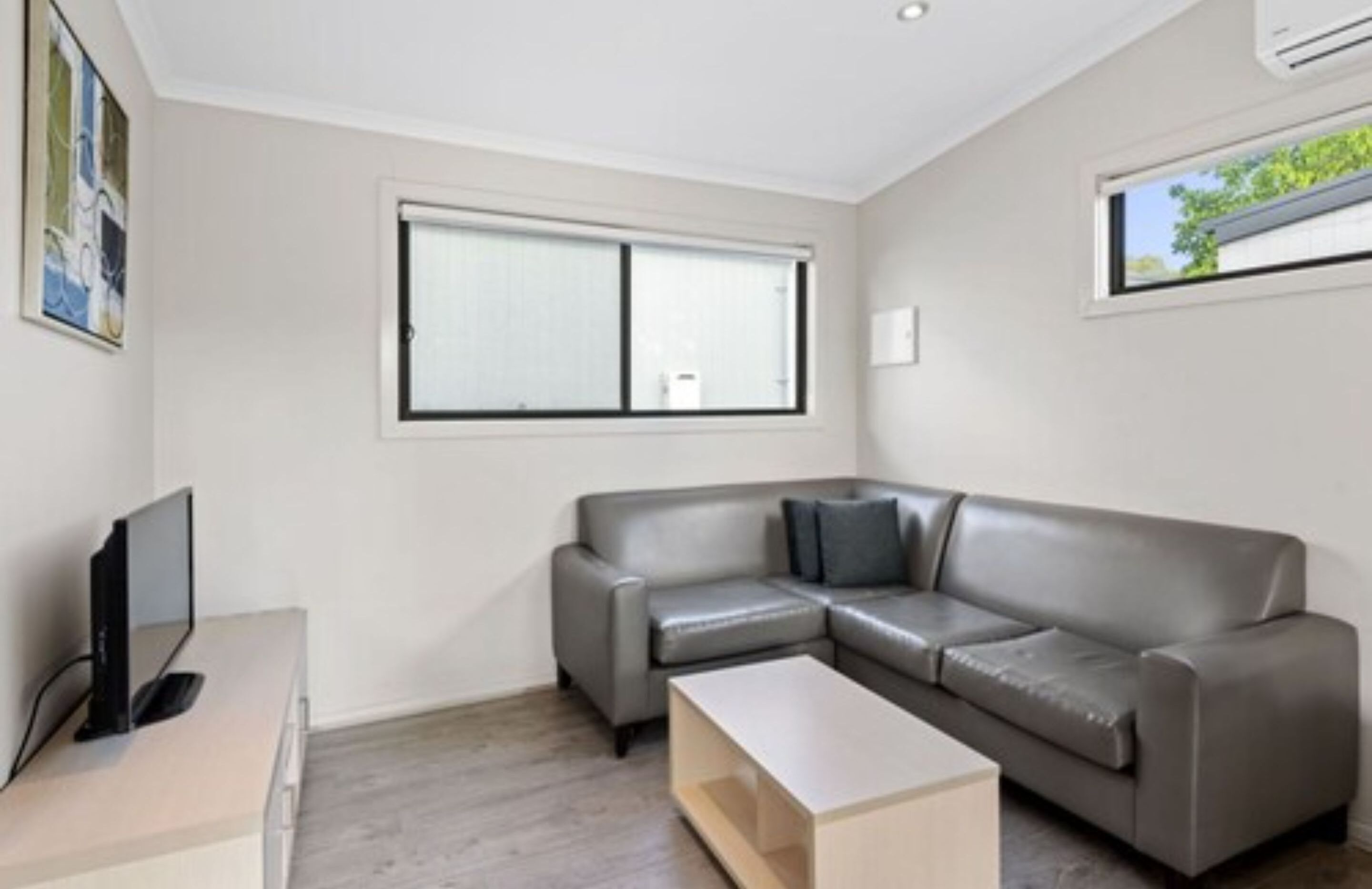 A modern living room with a grey leather sofa, a flat-screen TV, and a window with blinds.