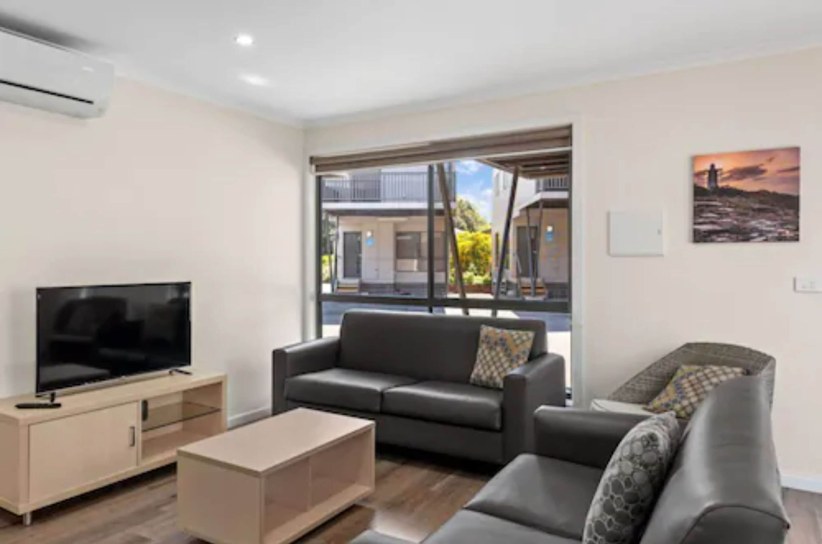 A modern living room with a flat-screen TV, a sofa, a coffee table, and a window offering a view of buildings and greenery.