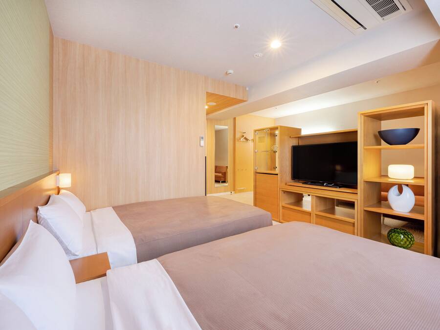 Quarto de hotel com duas camas, uma televisão, um armário de madeira e um banheiro visível por uma porta aberta.
