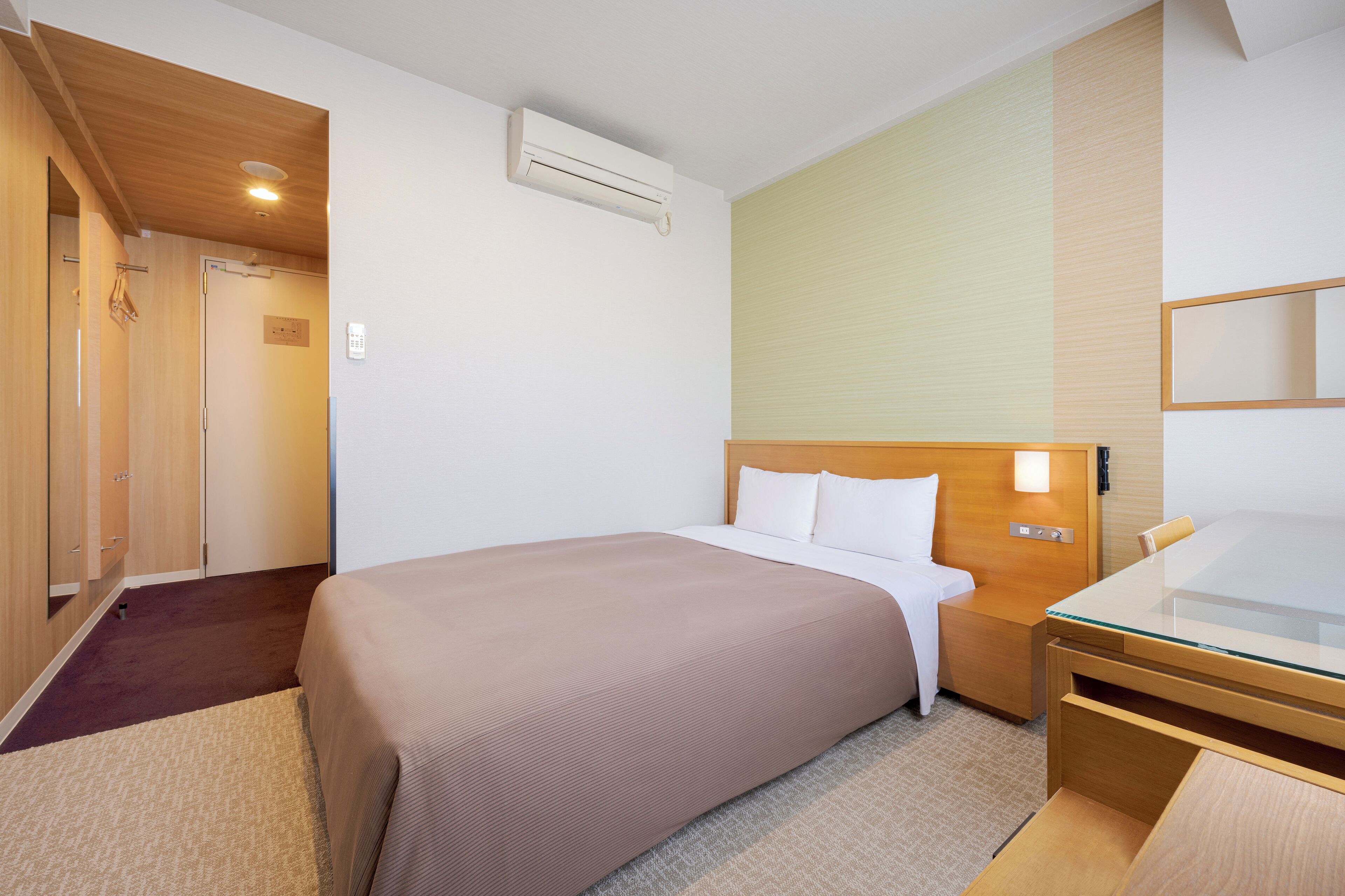 Quarto de hotel com cama, mesa de cabeceira e escrivaninha com tampo de vidro.