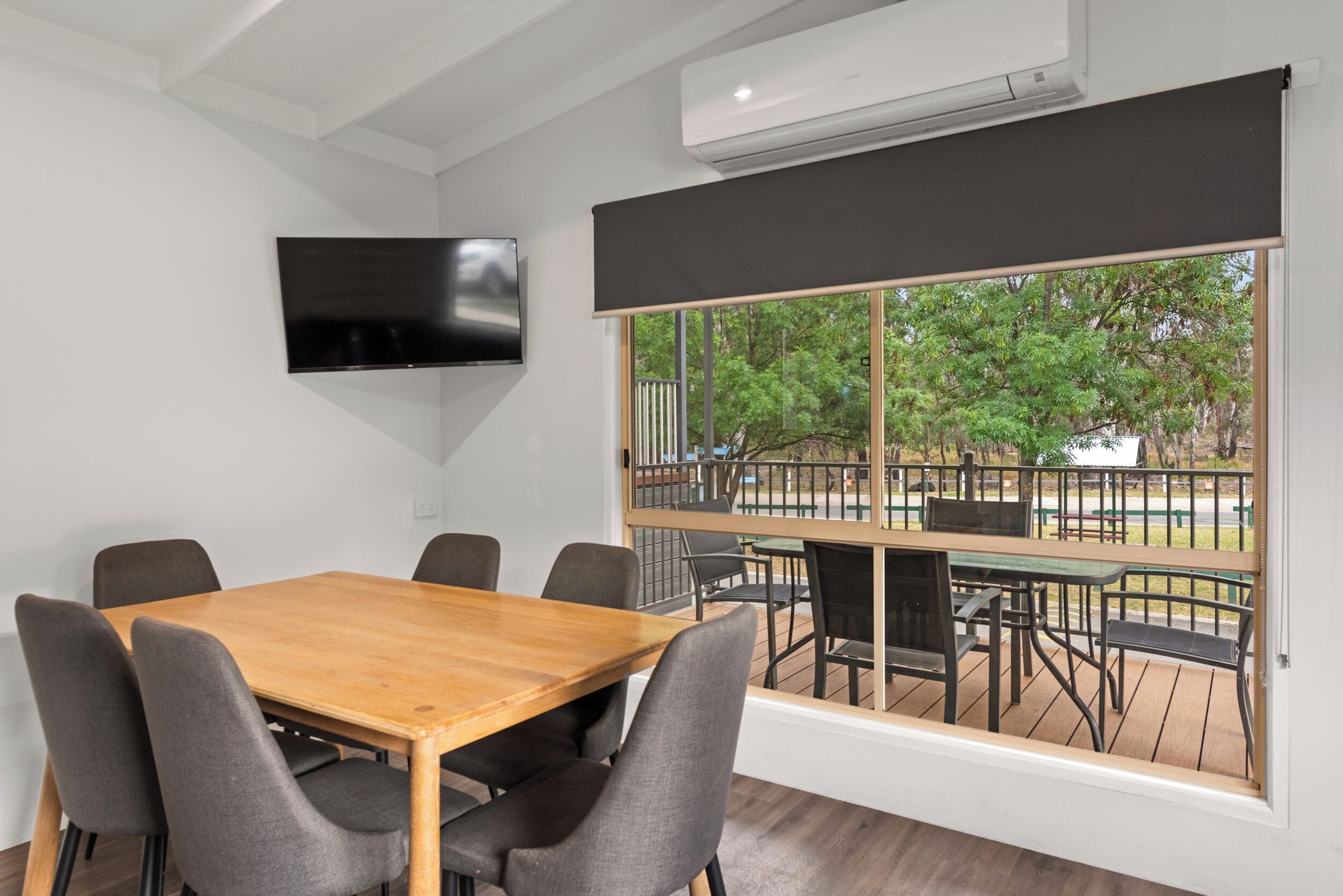 A conference room with a wooden table and grey chairs, a flat-screen TV mounted on the wall, and a large window overlooking a deck with outdoor furniture.