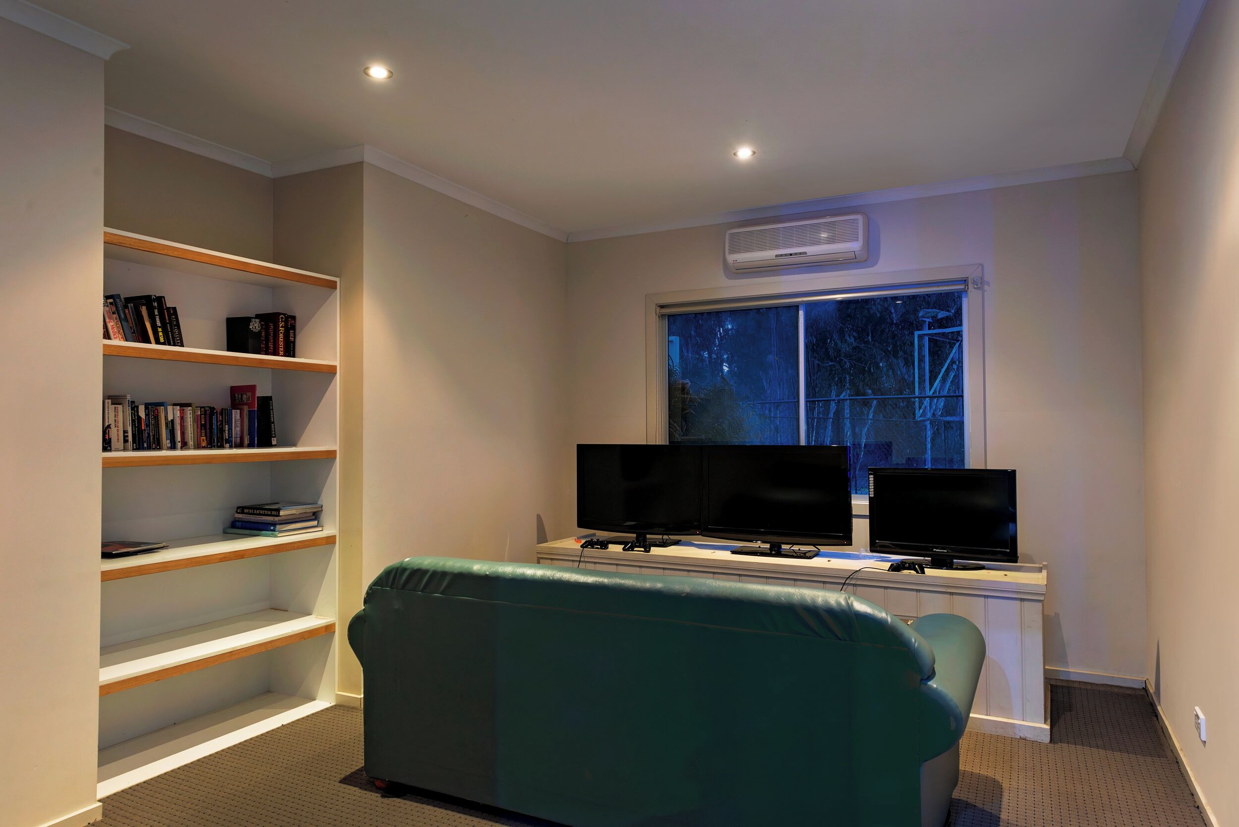 Una habitación con un escritorio, dos monitores, una estantería y un sofá verde.