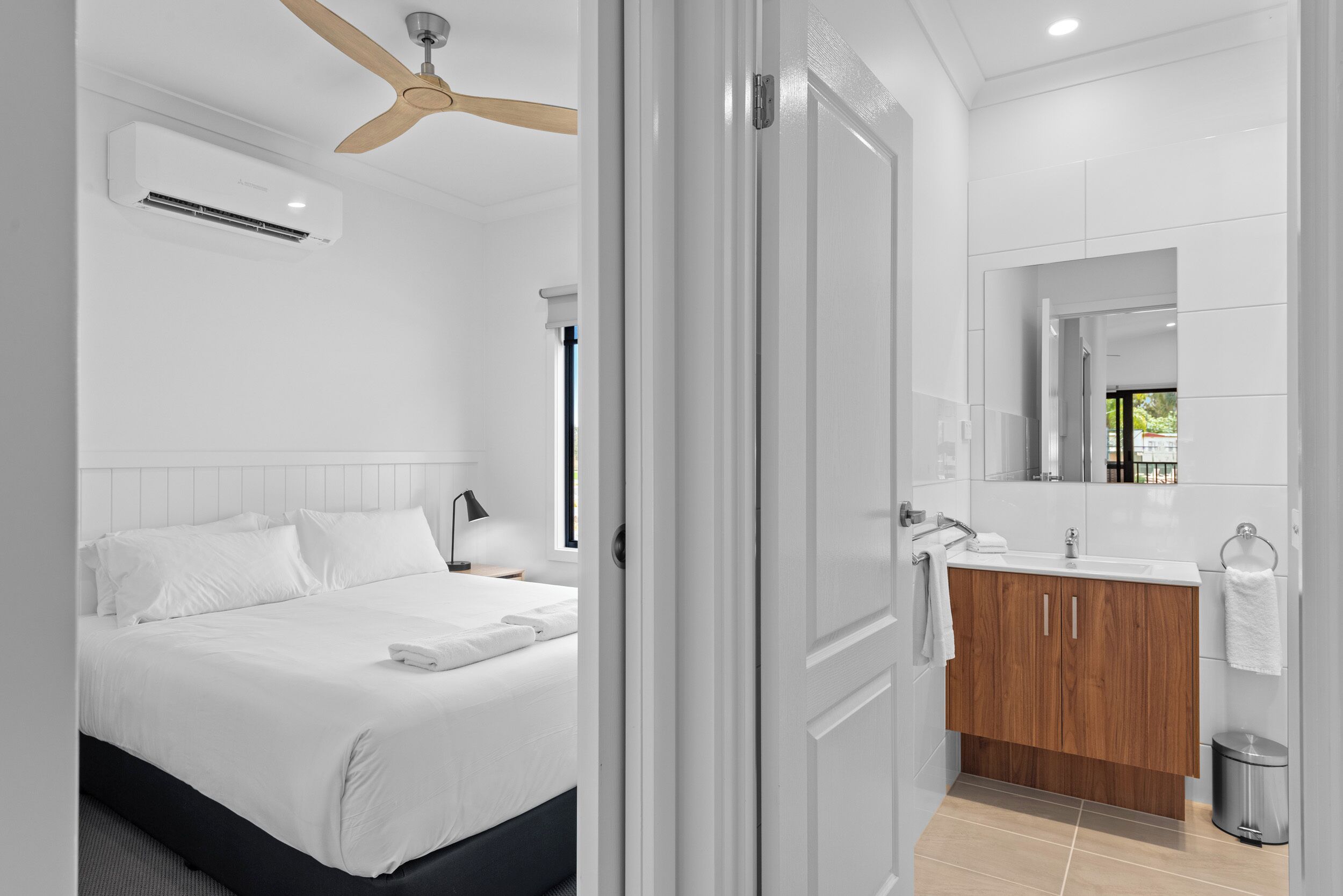 Een moderne hotelkamer met een bed, een plafondventilator van hout, een badkamer met een houten wastafel en een grote spiegel.