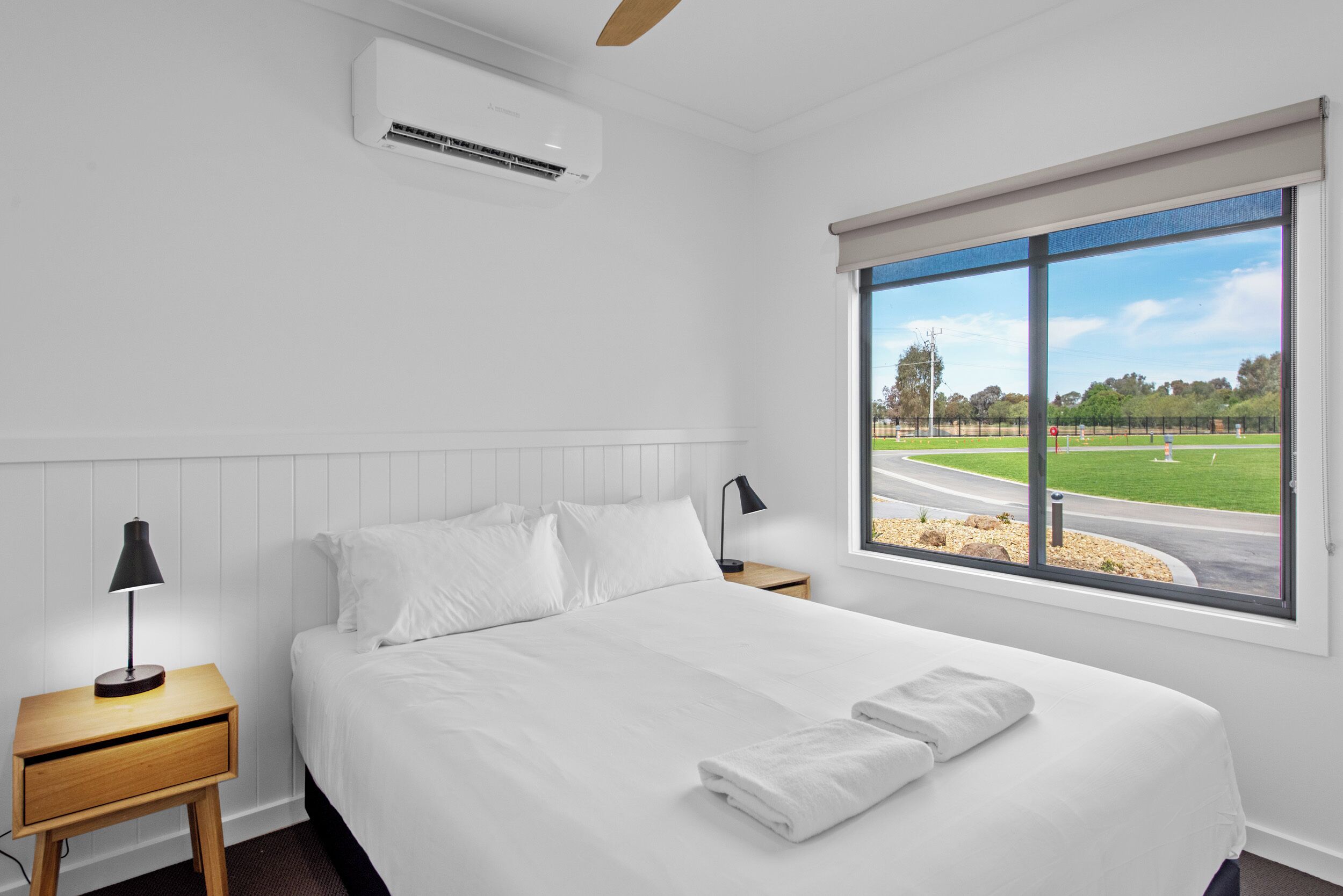 Habitación de hotel con una cama grande, una mesita de noche con lámpara, un equipo de aire acondicionado y una ventana con vistas a un parque y una carretera.