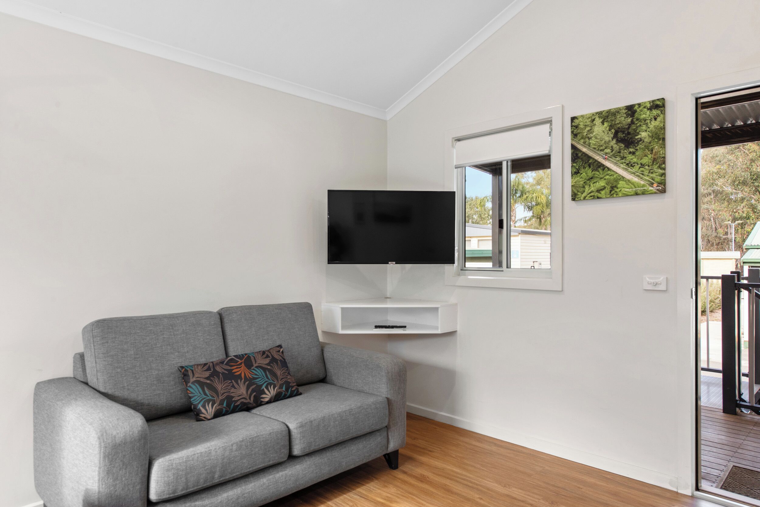 A modern living room with a grey sofa, a mounted TV, and a window with a view of greenery.