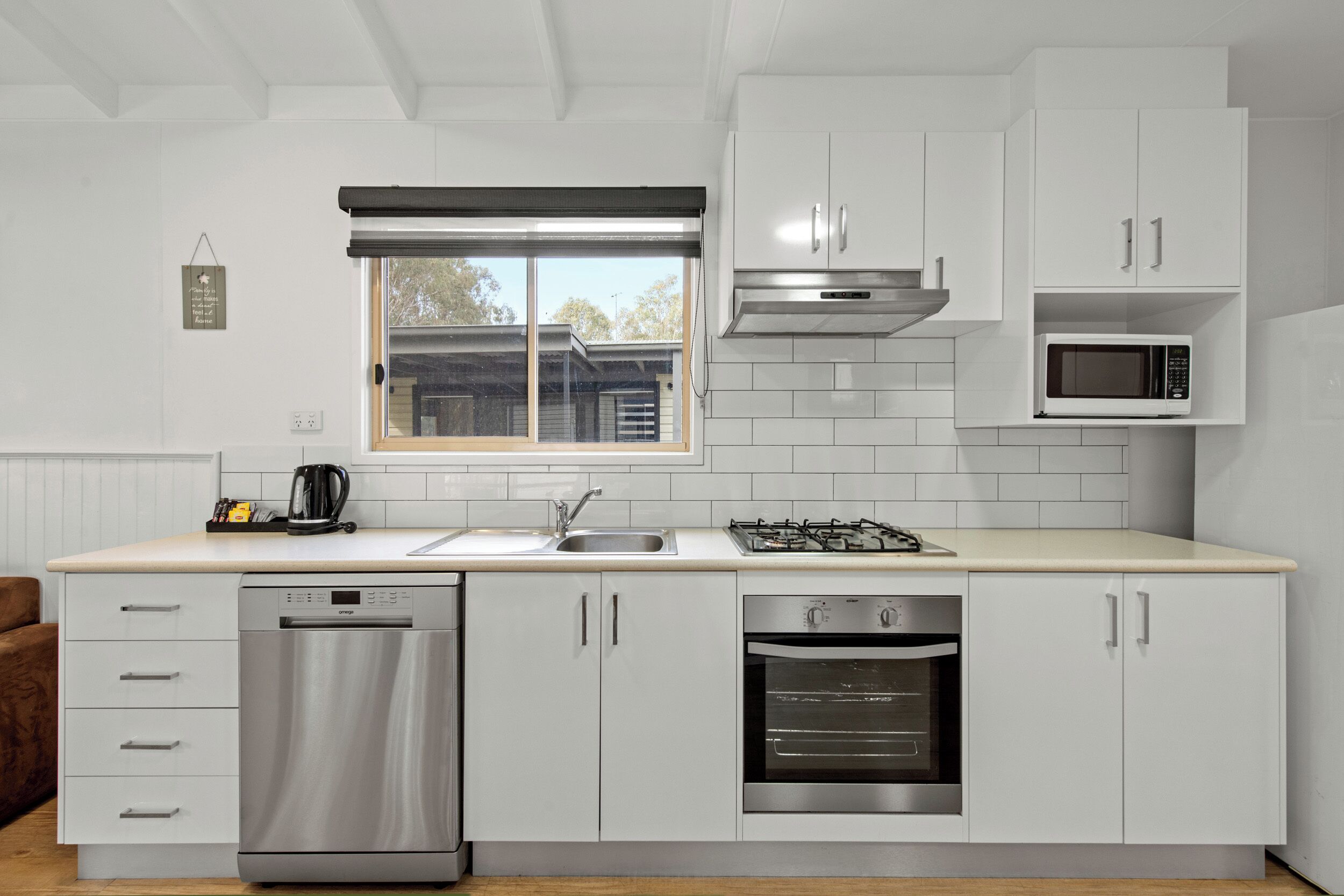 A modern kitchen with white cabinetry, a dishwasher, an oven, a microwave, and a window with a view of the outside.
