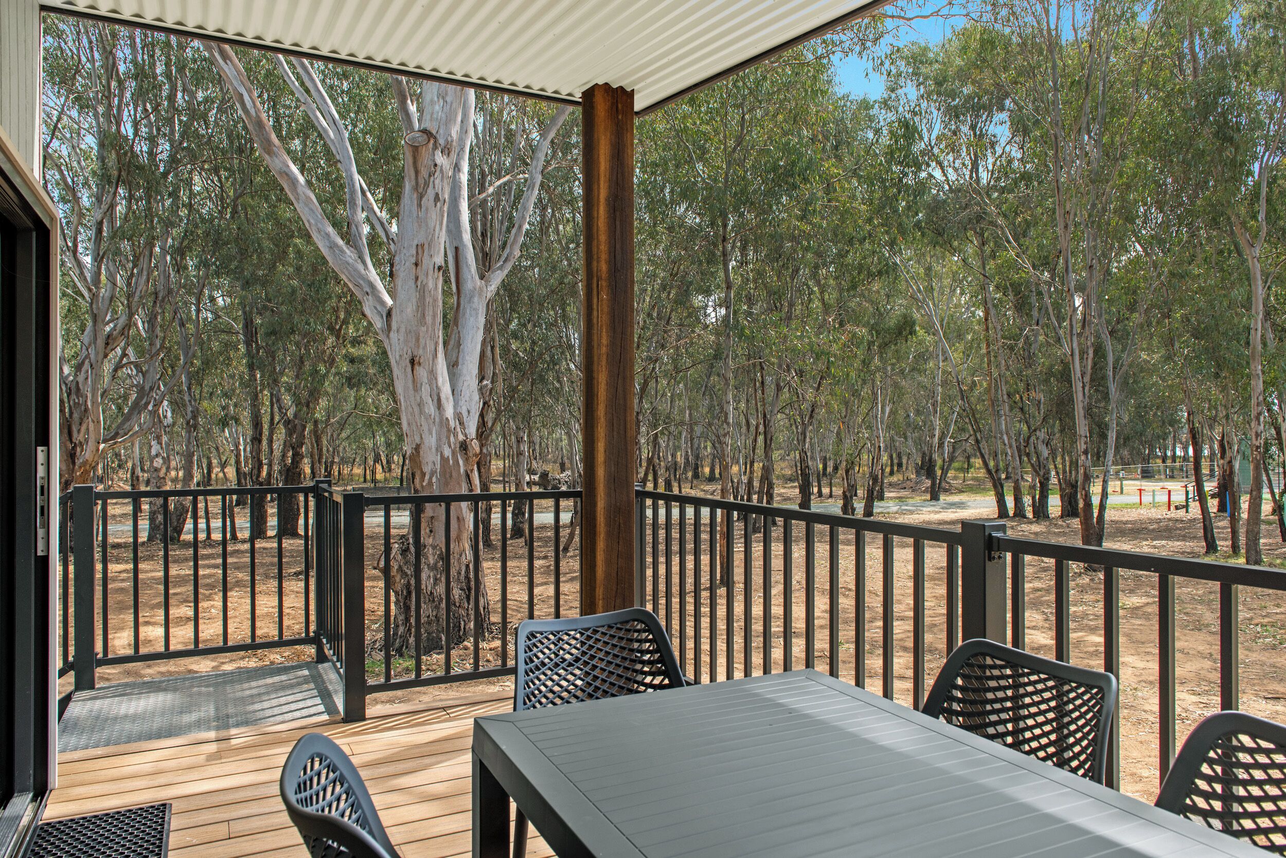 A covered outdoor area with a table and chairs, surrounded by a metal railing, overlooking a wooded area with trees and a dirt path.