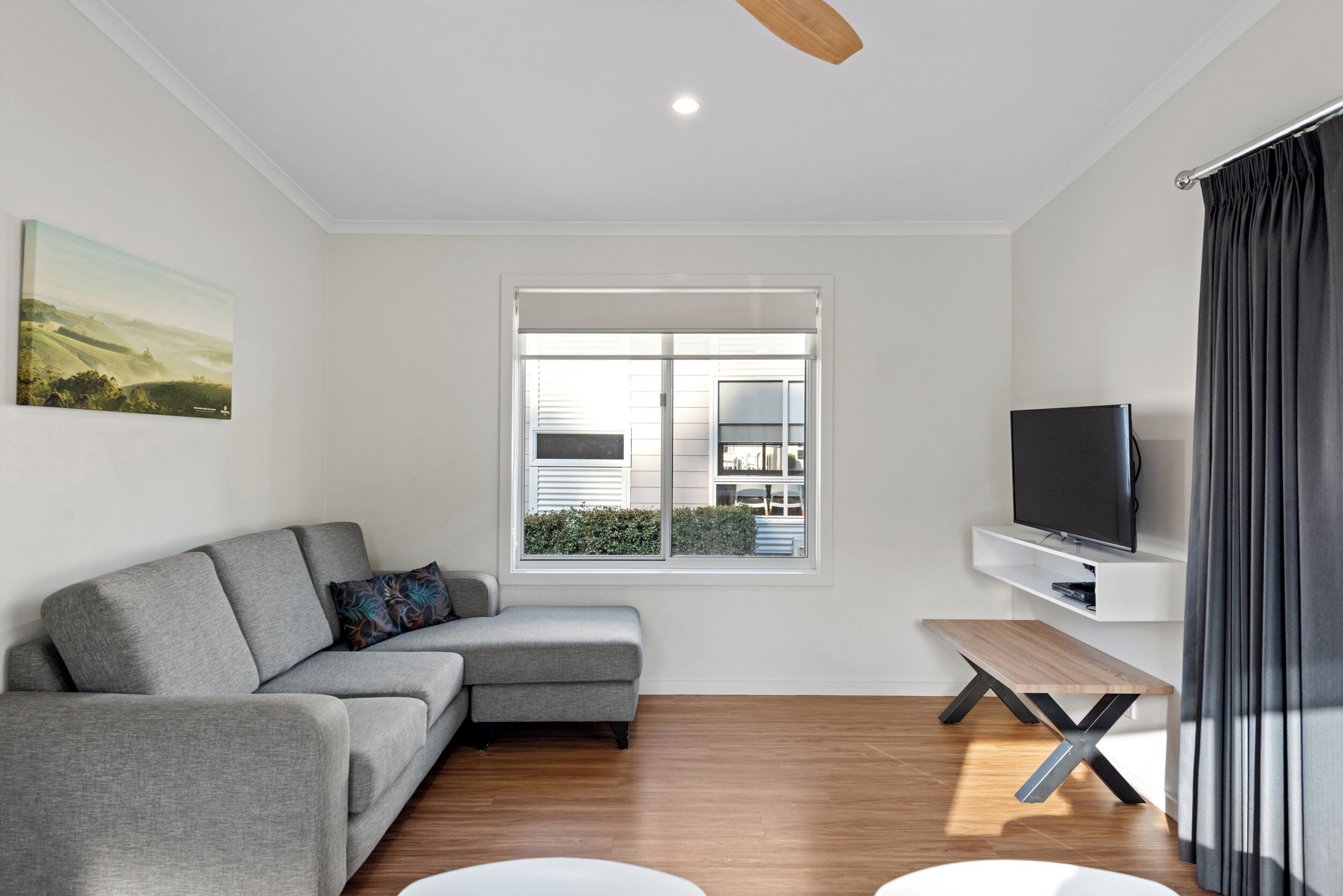 A modern living room with a grey sectional sofa, a wooden coffee table, a flat-screen TV, and a large window with blinds.
