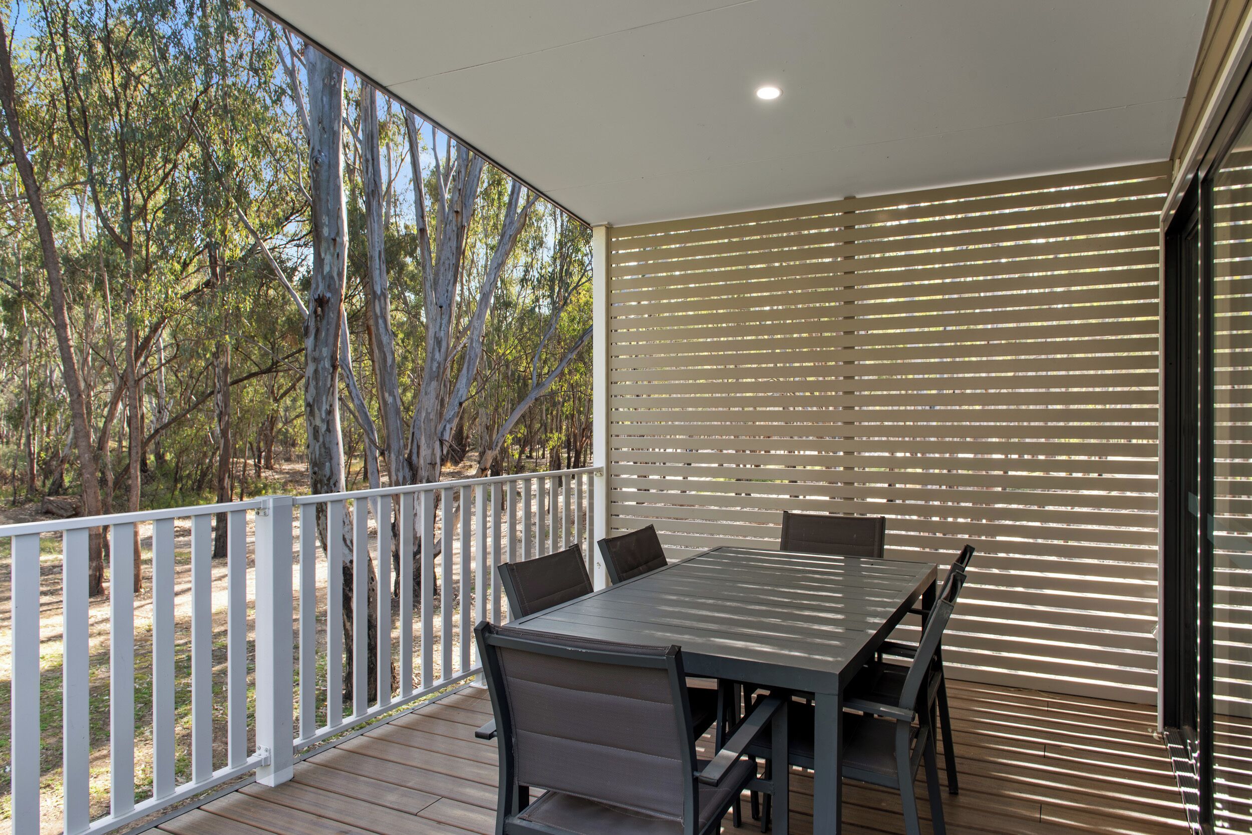 A covered outdoor patio with a table and chairs, surrounded by a white fence and trees.