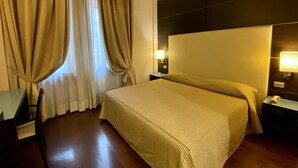 Una habitación de hotel con una cama grande, dos mesitas de noche con lámparas, piso de madera y cortinas color beige.