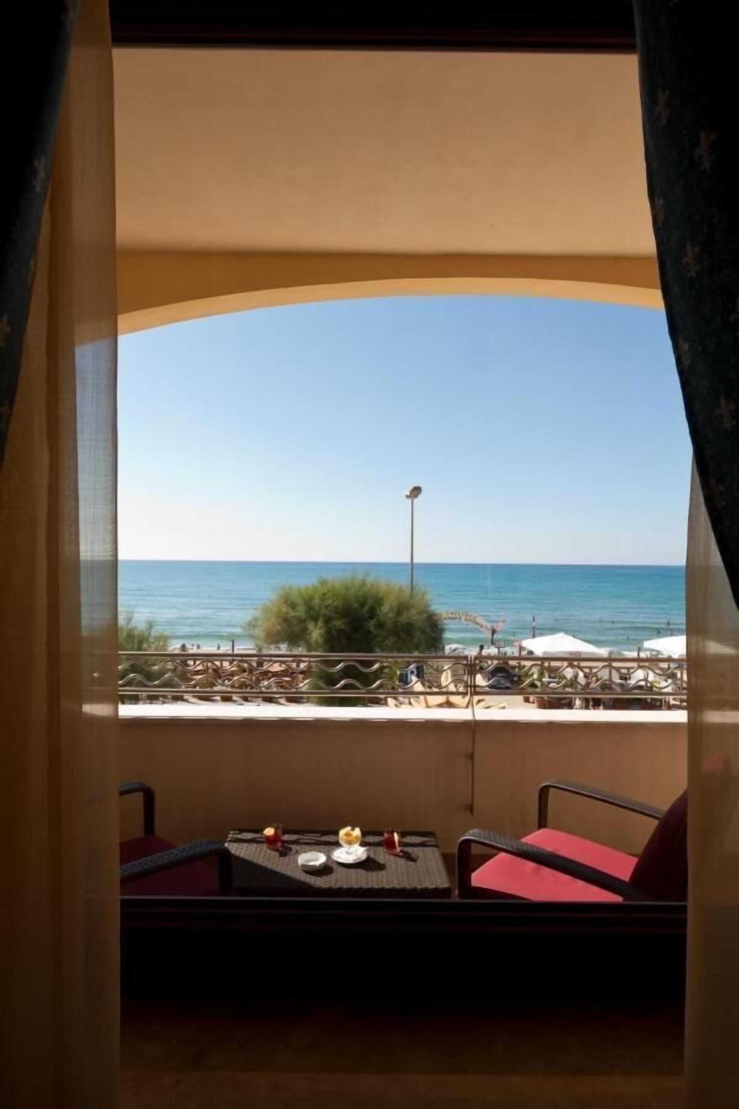 A balcony with a table and chairs, overlooking a beach with umbrellas and people.