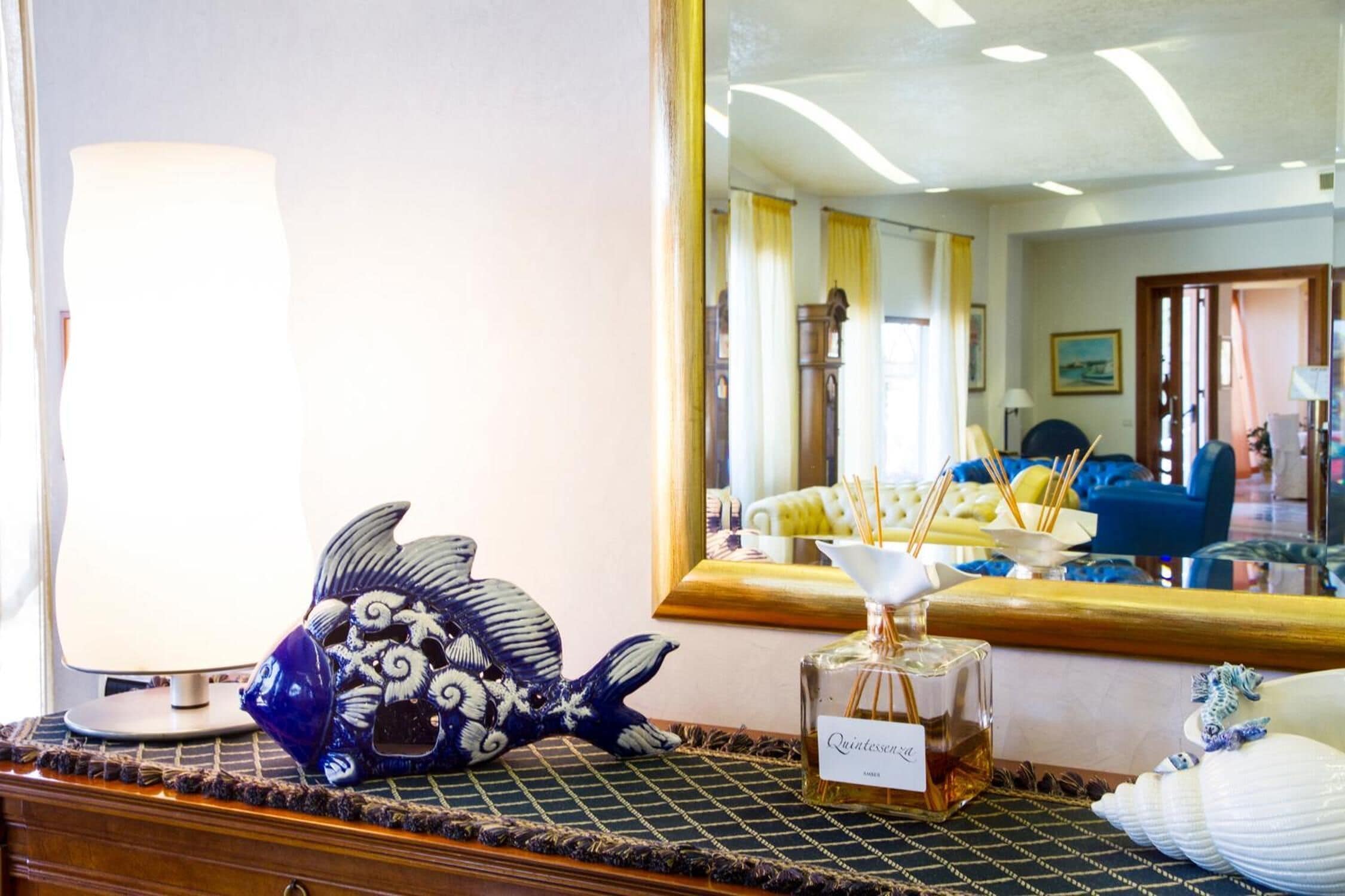 A room with a large mirror reflecting a yellow sofa and a wooden cabinet. A decorative fish sculpture and a bottle of Quiletteson are on a table.