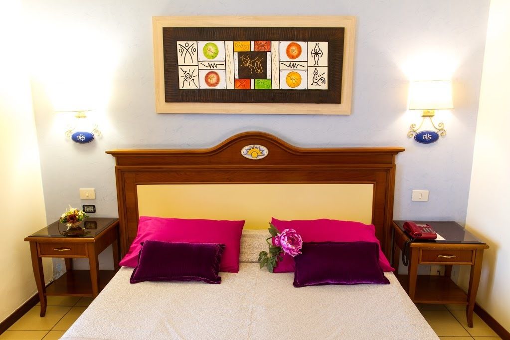 A double bed with two purple pillows, a wooden headboard, a framed artwork above, bedside tables with lamps, and a telephone on one table.