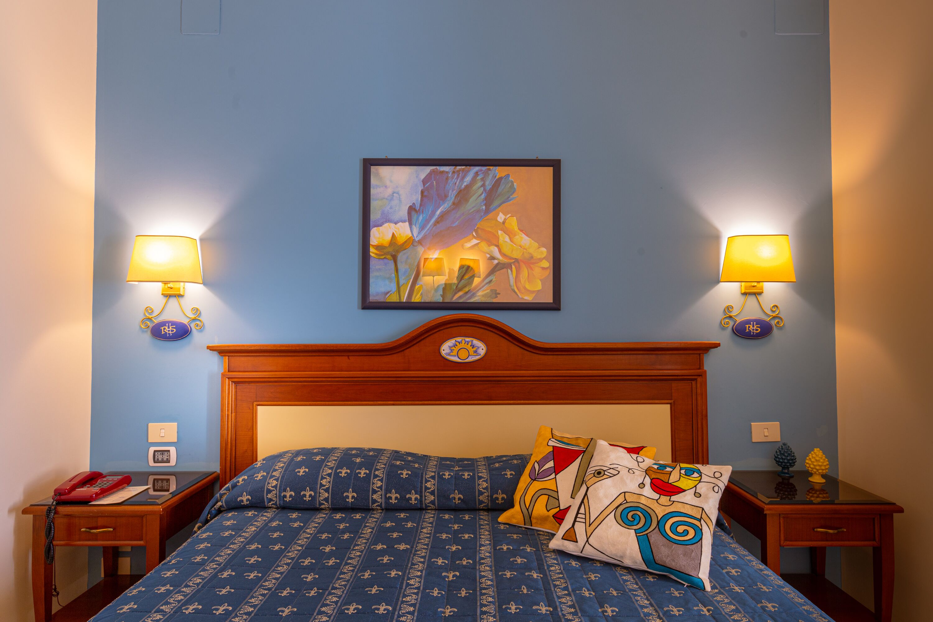 A bedroom with a bed, two bedside tables, a nightstand, a wall-mounted painting, and two wall-mounted lamps.