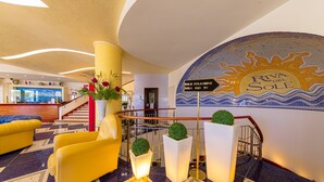 A hotel lobby with a reception desk, yellow armchairs, a staircase, and a mural of the sun. - Hotel Riva del Sole (Cefalù)