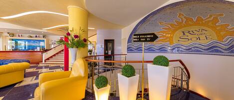 A hotel lobby with a reception desk, yellow armchairs, a staircase, and a mural of the sun.