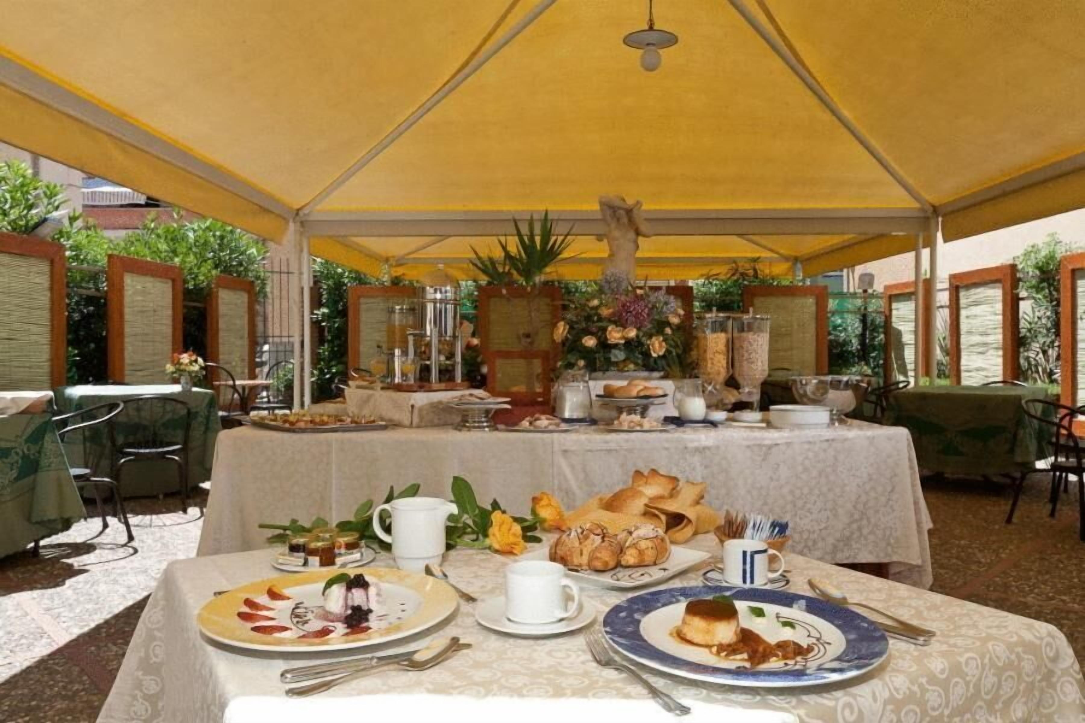 Zona pranzo riparata con tavoli apparecchiati per la colazione, con diversi piatti e un centrotavola floreale.