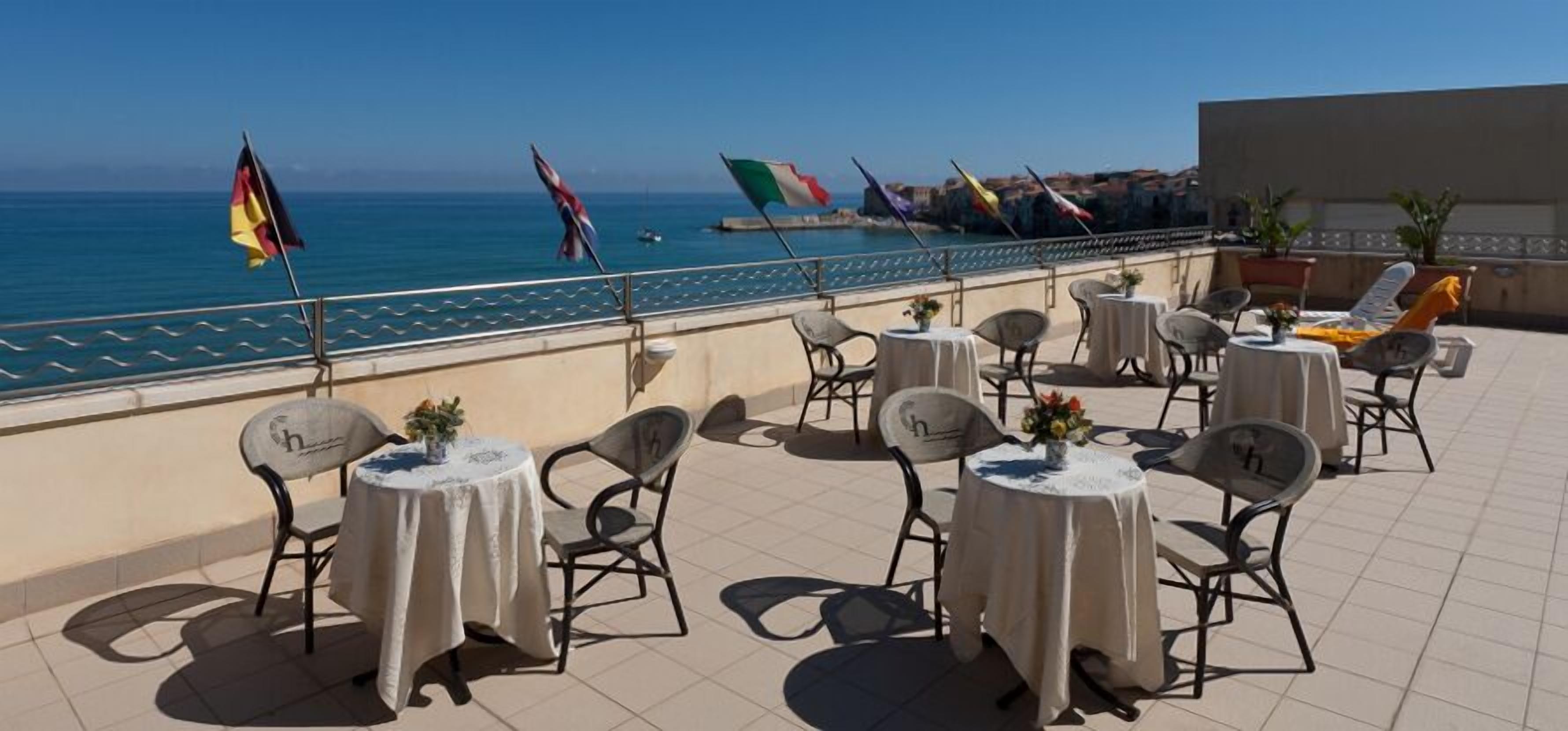 A terrace with round tables and chairs, overlooking a body of water with flags.