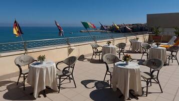Une terrasse avec des tables et des chaises rondes, donnant sur une étendue d’eau où flottent des drapeaux.