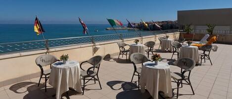 A terrace with round tables and chairs, overlooking a body of water with flags.