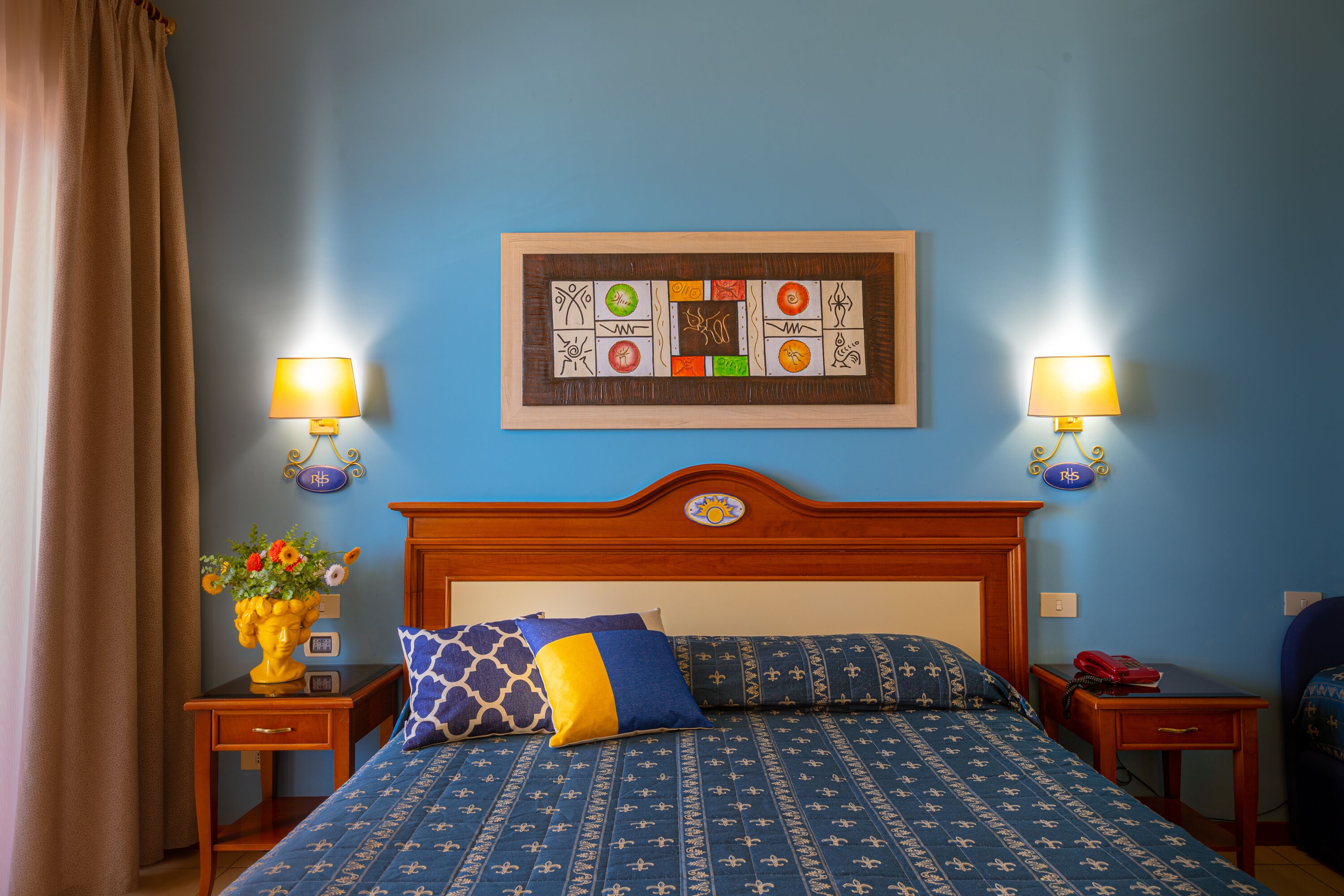 Habitación de hotel con cabecera de madera, colcha estampada azul, dos mesitas de noche con lámparas, un cuadro enmarcado sobre la cama y un jarrón con flores en la mesita de noche.