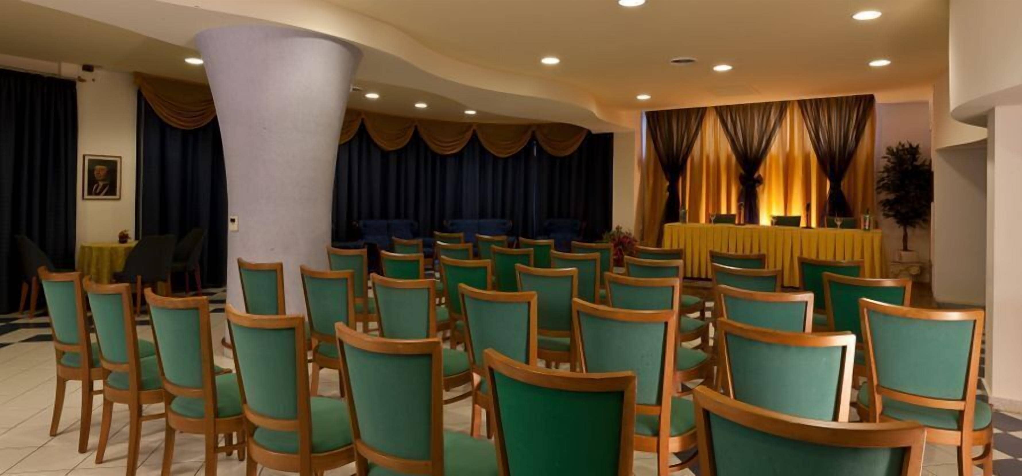 A conference room with rows of green chairs, a white pillar, and a stage with a yellow table.