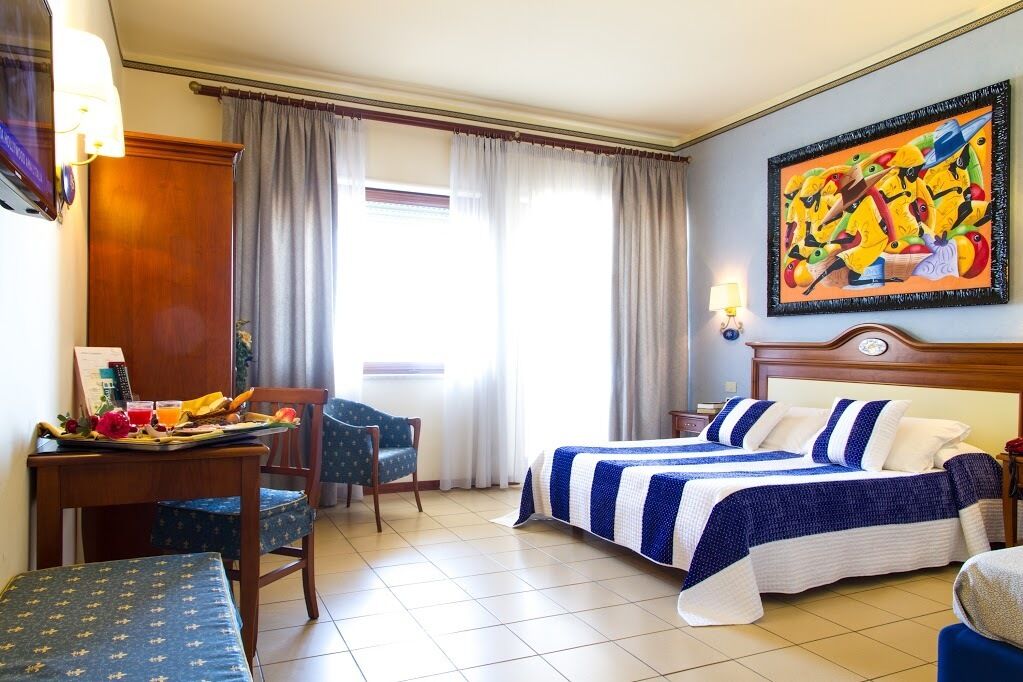 Ein Hotelzimmer mit zwei Betten, einem Esstisch mit Frühstücksangeboten, einem Stuhl und einem Bild an der Wand.