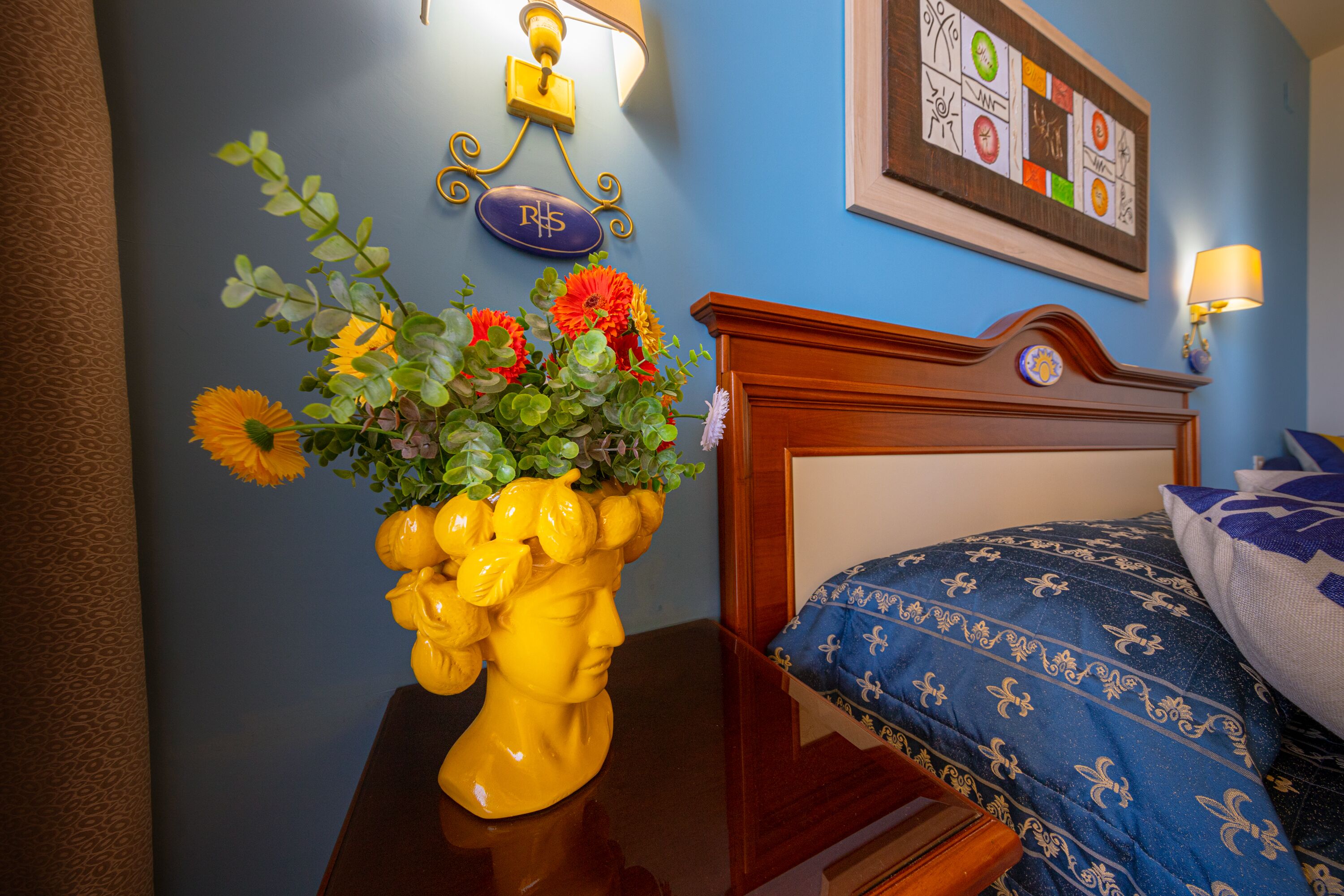 A hotel room with a wooden headboard, a bed with blue bedding, a vase with flowers, and a wall-mounted sign with the initials "RHS".