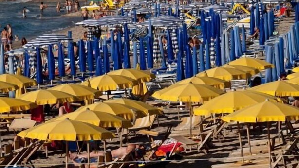 A beach with rows of sun umbrellas and beach chairs, a castle in the background, and a clear blue sky. - Hotel Riva del Sole (Cefalù)