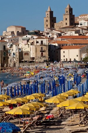 A beach with rows of sun umbrellas and beach chairs, a castle in the background, and a clear blue sky. - Hotel Riva del Sole (Cefalù)