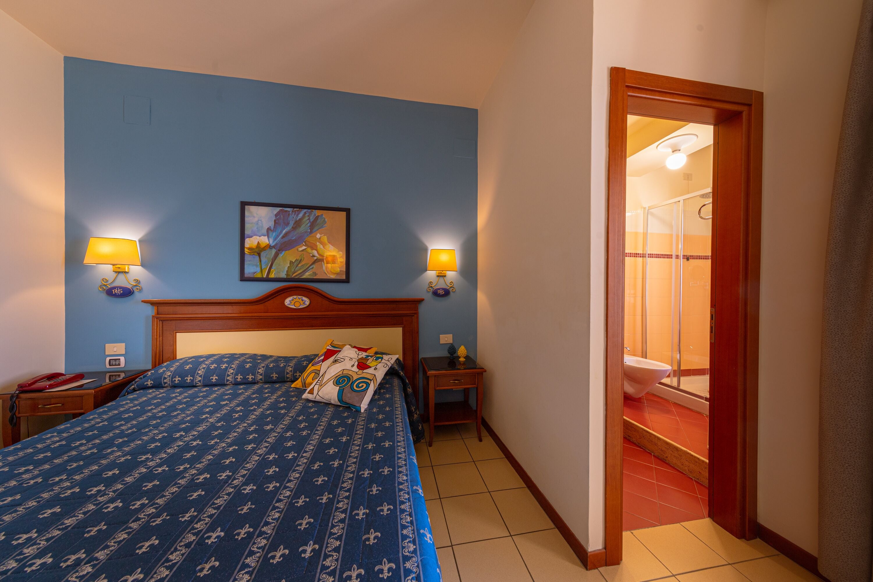 A hotel room with a bed, two bedside lamps, a painting, a nightstand, and a bathroom visible through an open door.