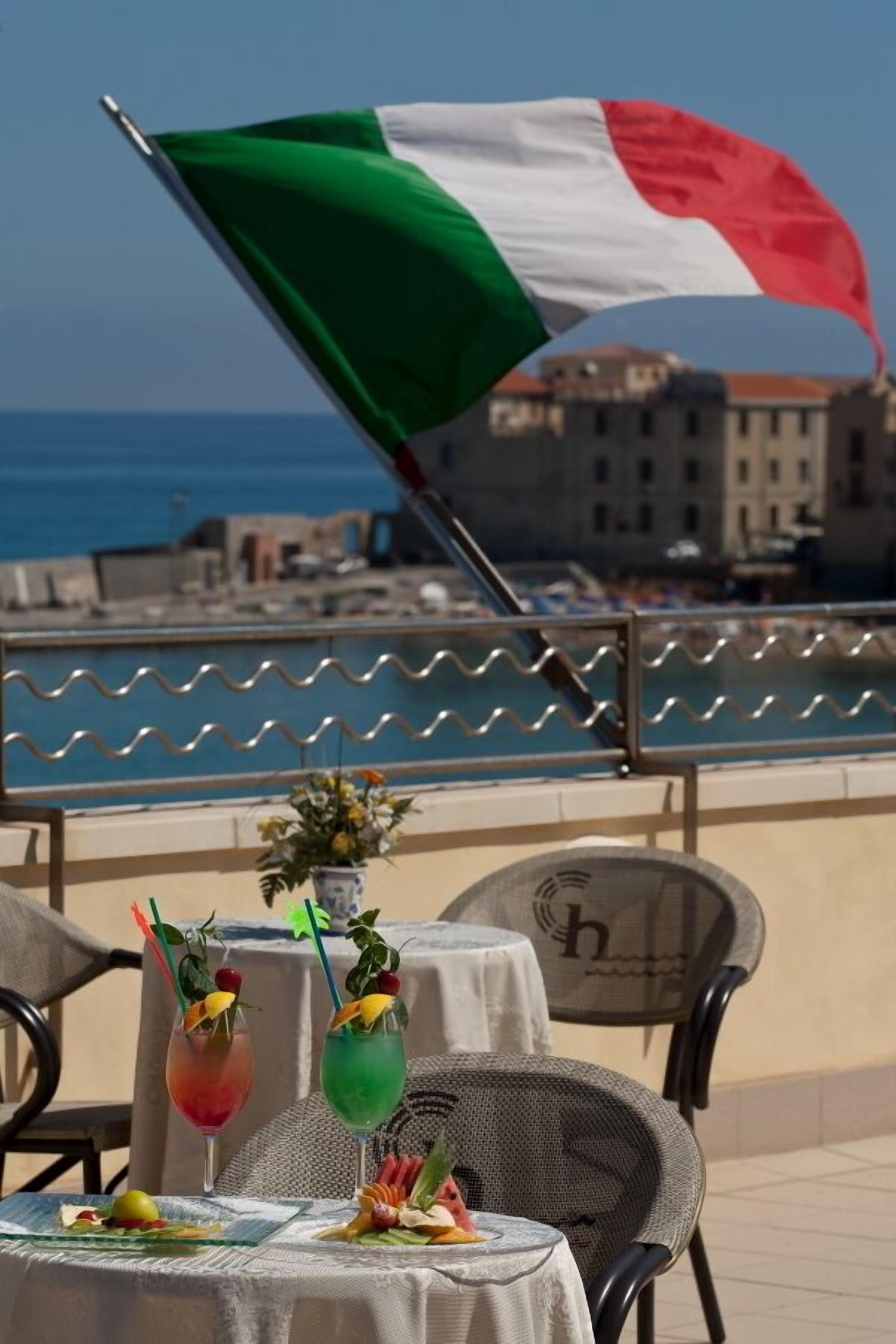 A table with drinks and a flag, overlooking a coastal area with buildings and the sea.