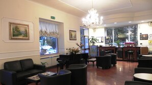 A hotel lobby with black leather sofas, a piano, a chandelier, and a bar area with a view of the street outside. - Villa Robinia (Genzano di Roma)