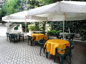 Eine Terrasse im Freien mit Tischen und Stühlen, beschattet durch Sonnenschirme und umgeben von Grünpflanzen.