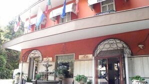 A building with a red facade, a fountain, and multiple flags. - Villa Robinia (Genzano di Roma)