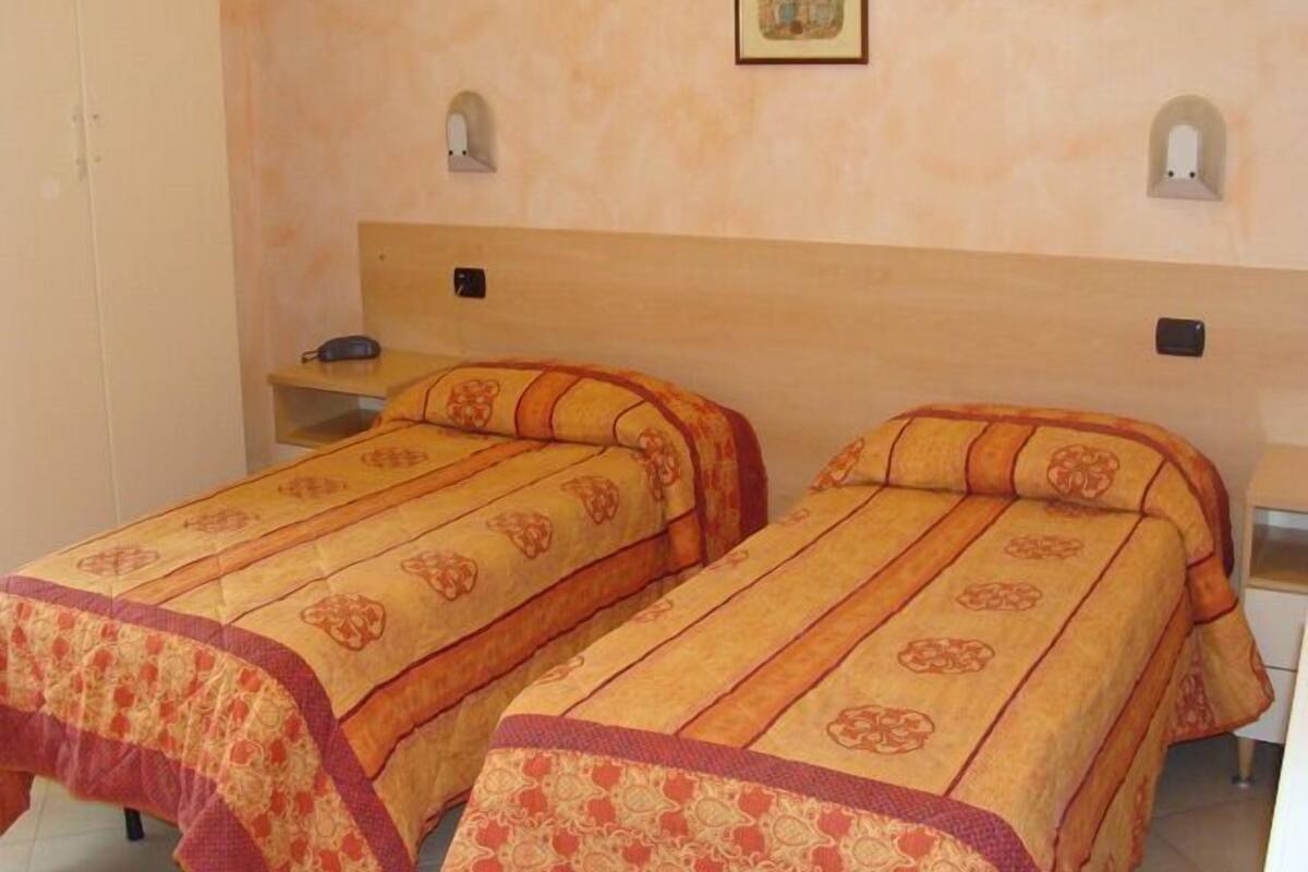 Une chambre avec deux lits simples, chacun agrémenté d’une couvre-lit et de coussins à motifs, d’une petite étagère et d’un tableau encadré au mur.