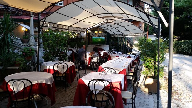 An outdoor restaurant with tables covered by canopies, surrounded by potted plants and a TV screen. - Villa Robinia (Genzano di Roma)