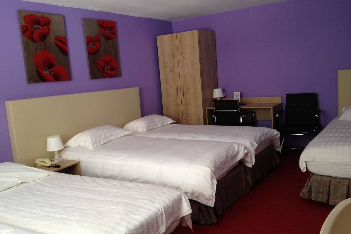 Une chambre d’hôtel avec trois lits, une armoire en bois, un bureau avec une chaise et deux tableaux représentant des fleurs rouges accrochés au mur.