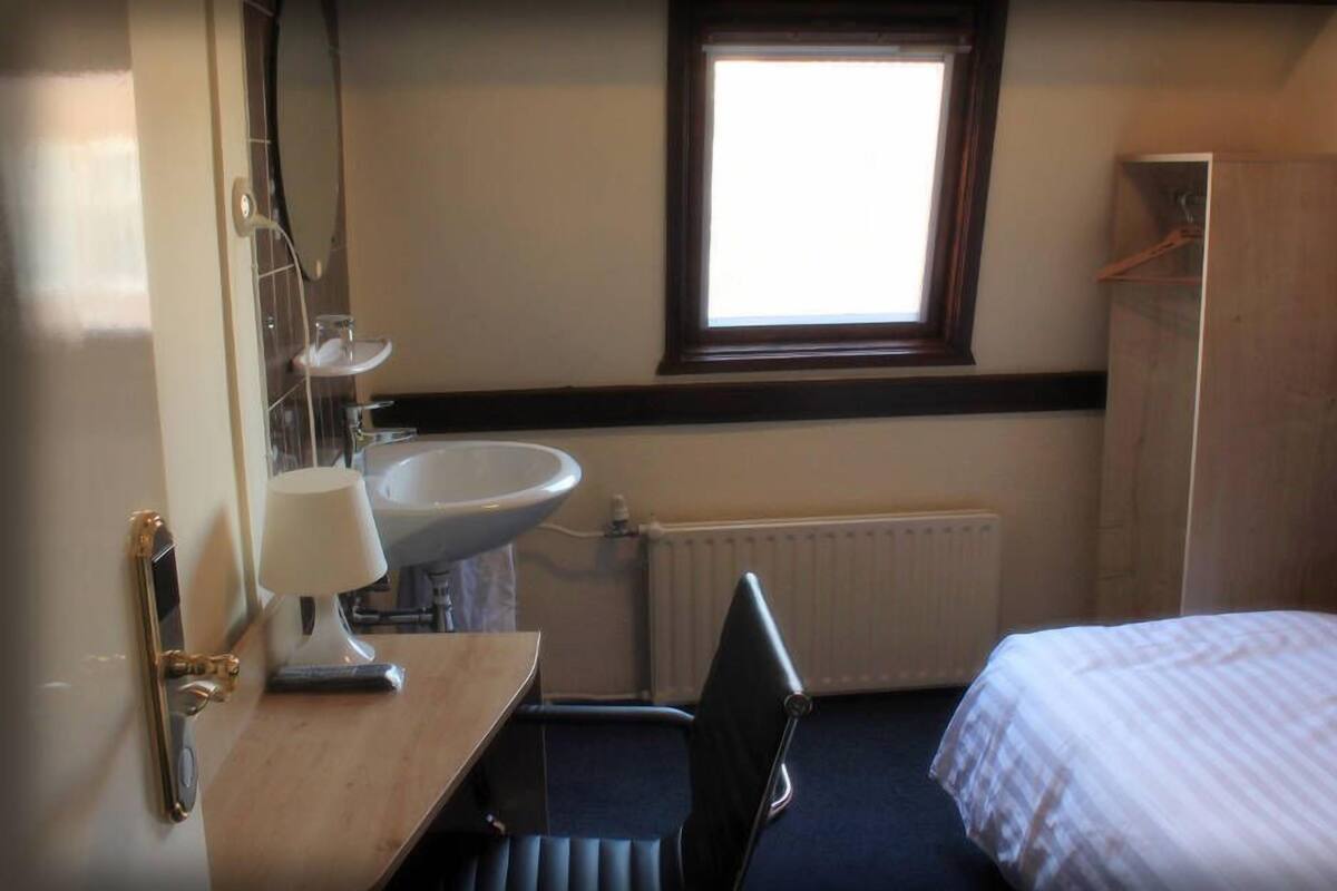 Une petite chambre d’hôtel, de taille réduite, comprenant un lit simple, une table de chevet avec une lampe, un lavabo et une fenêtre laissant entrer la lumière naturelle.