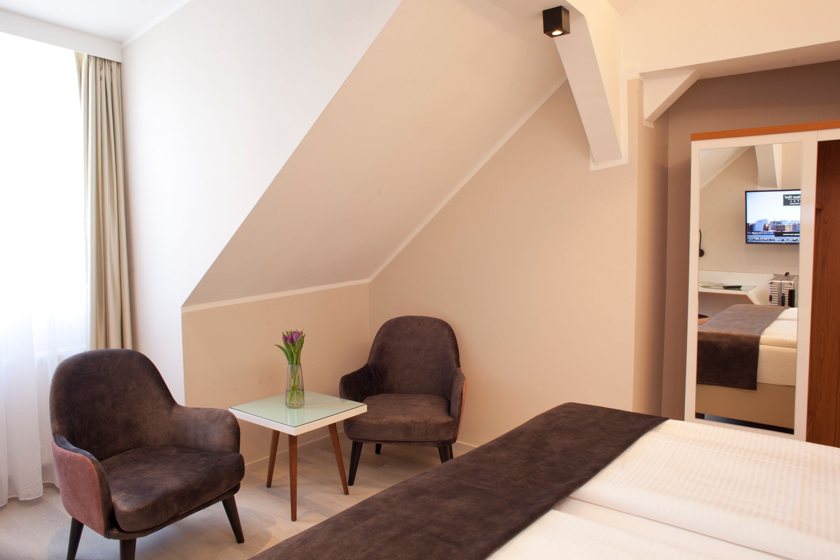Habitació d'hotel amb un llit, dues butaques, una taula petita amb un vas i una televisió penjada a la paret.