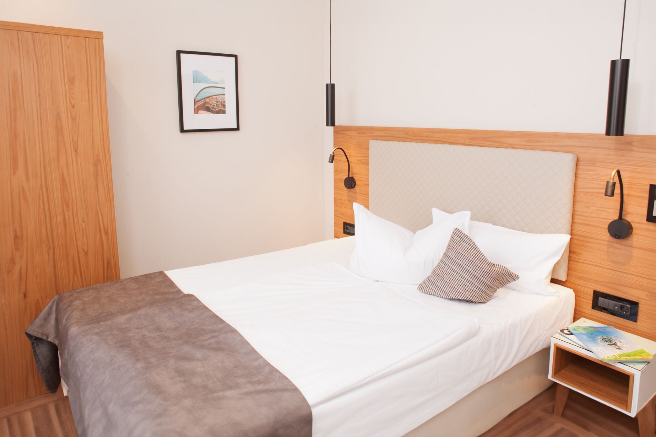 Un lit bien fait, avec des draps blancs et un plaid marron, une tête de lit en bois, une petite table de chevet avec un magazine et un tableau encadré accroché au mur.