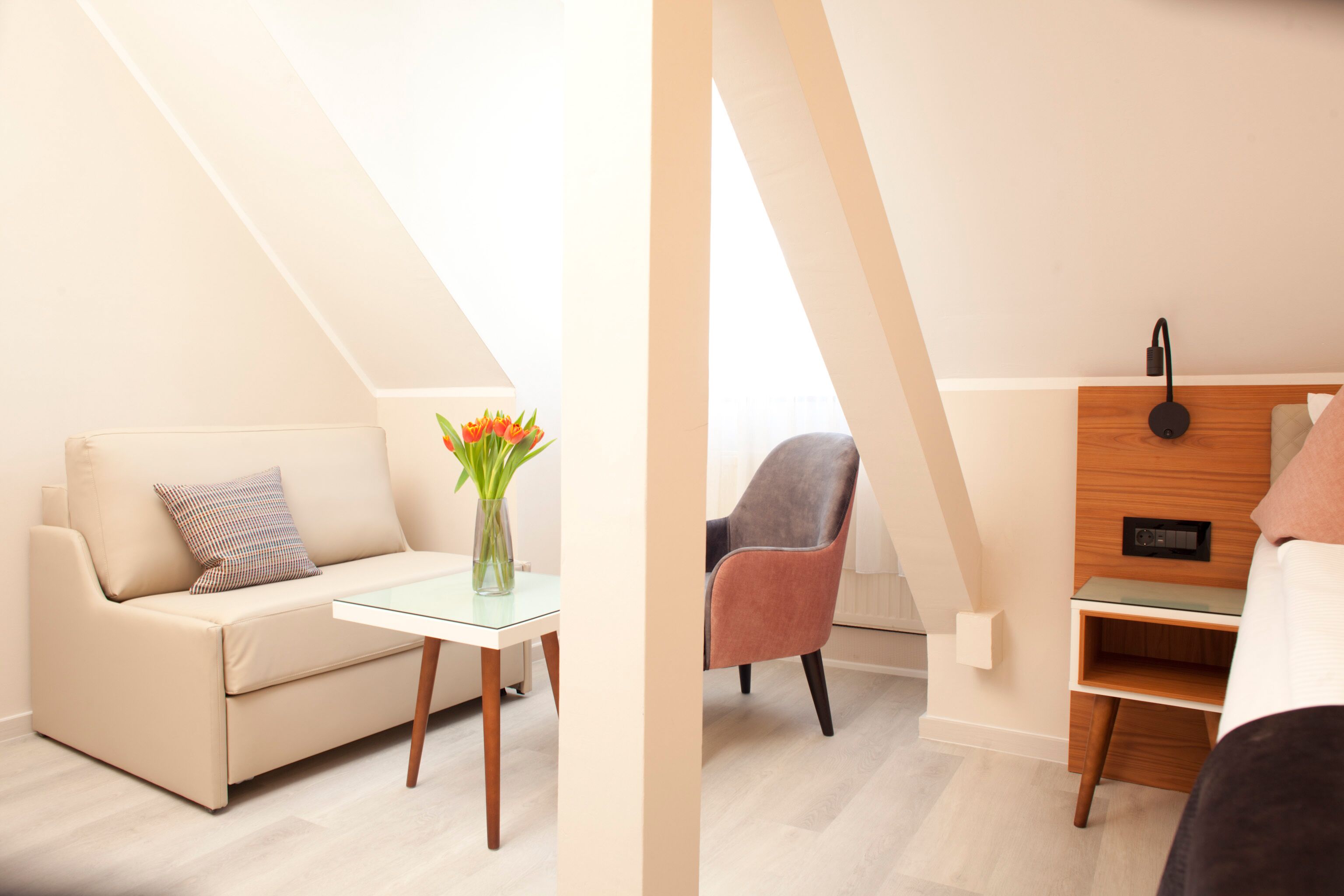 Una habitación moderna y luminosa con sofá, una mesita con un florero, una silla y una mesita de noche de madera con lámpara.