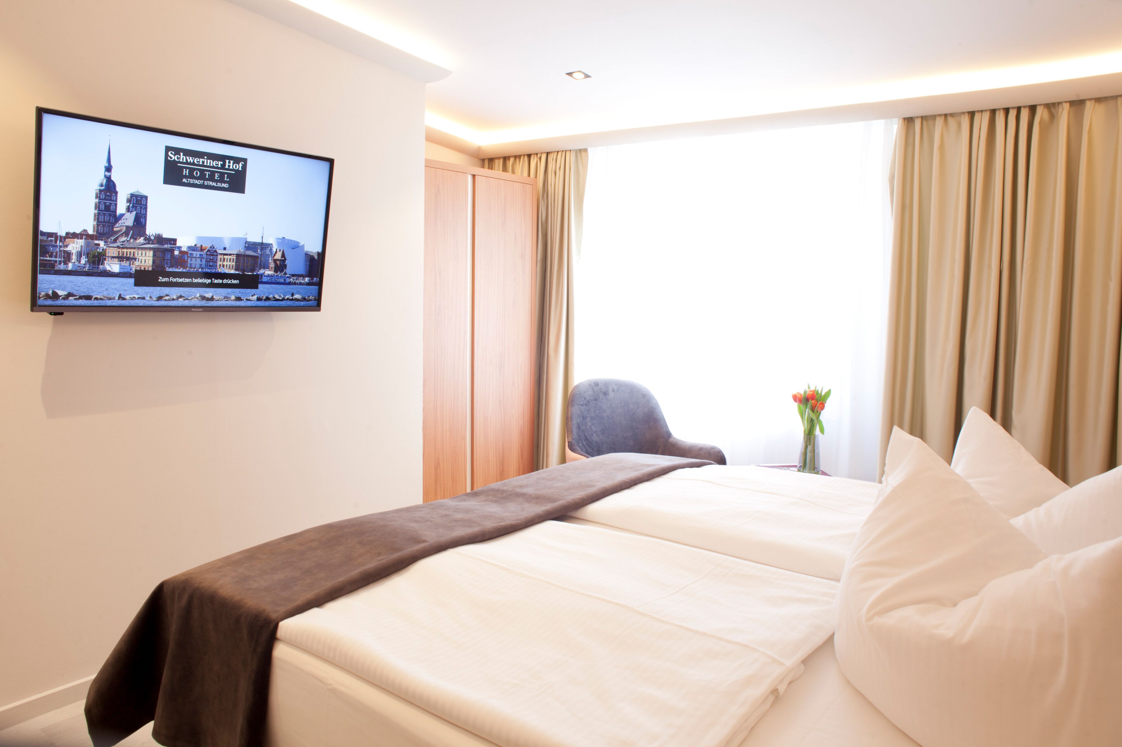 A hotel room with two beds, a TV displaying a cityscape, a desk with a chair, and a window with curtains.