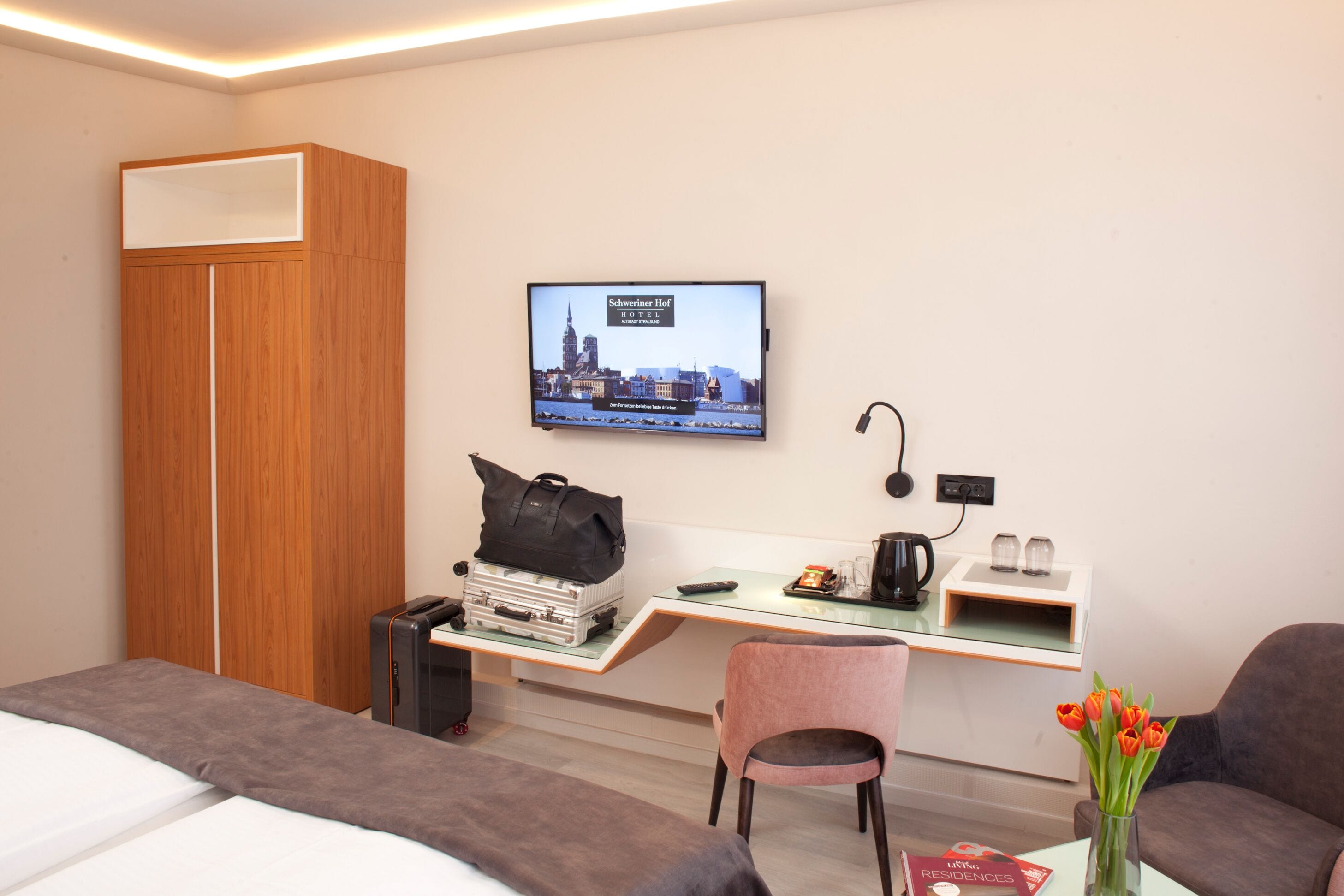 Habitación de hotel con una cama, un escritorio, una silla, un televisor que muestra una vista urbana y un florero con flores.